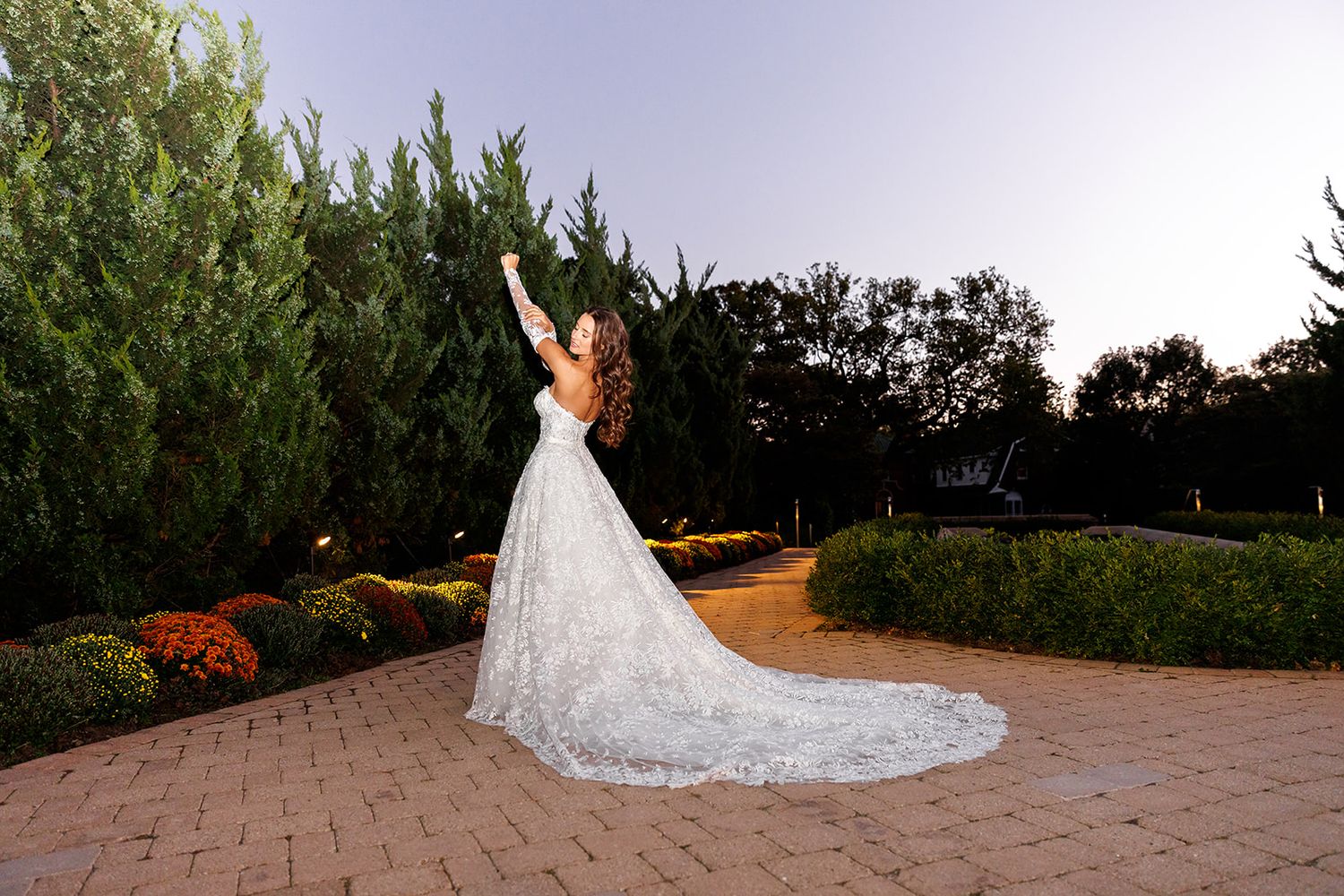 Bride in lace wedding gown raises arms joyfully on brick path surrounded by lush greenery at dusk.