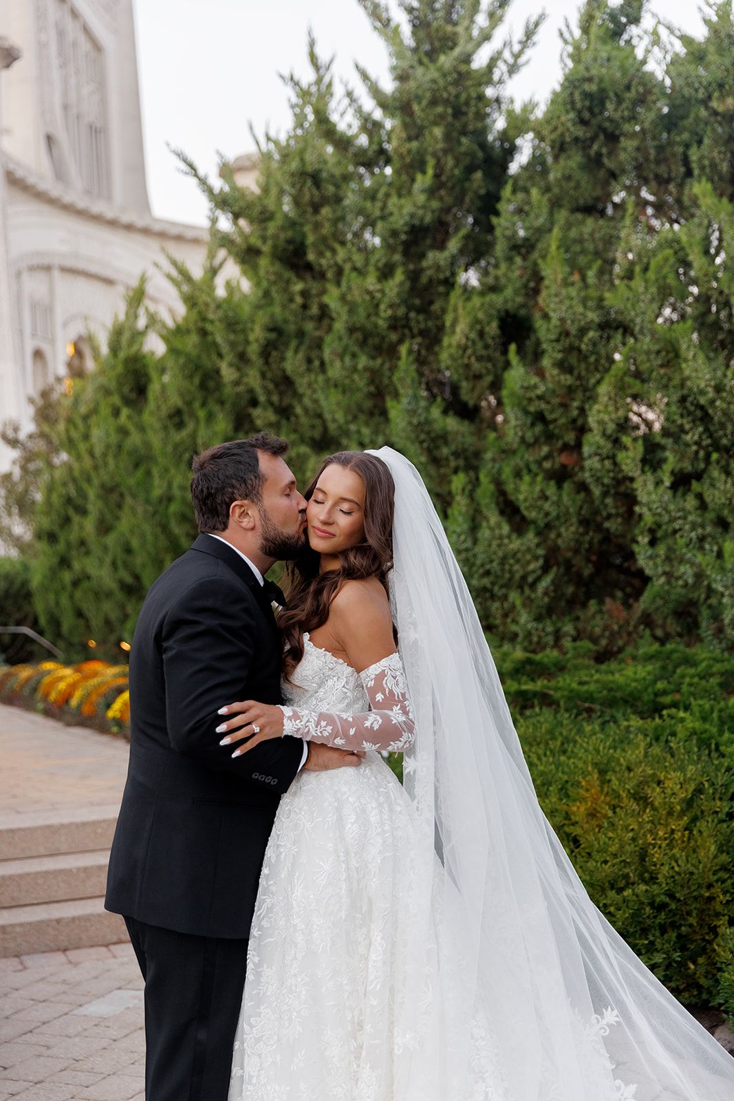 Groom tenderly kisses bride on the cheek beside lush green trees, bride in elegant off-shoulder lace gown and cathedral veil.