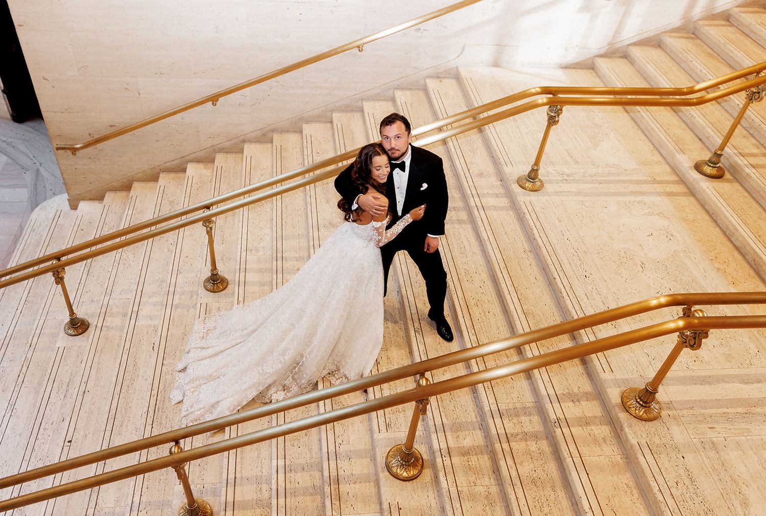Bride and groom pose on elegant marble staircase with gold railings at wedding venue.