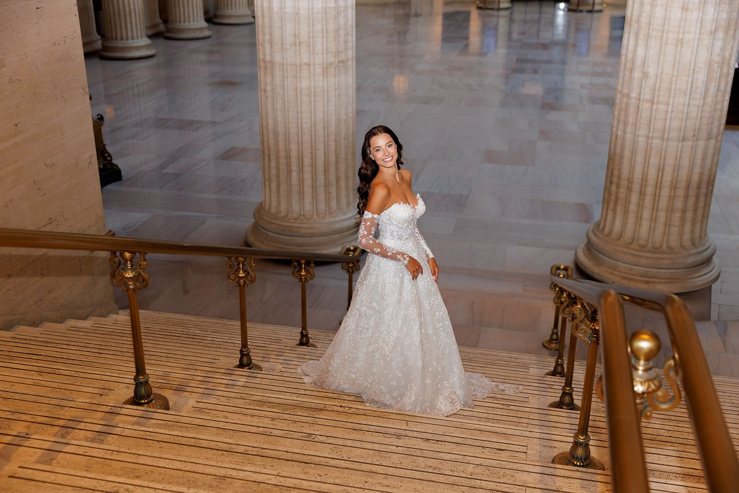 Elegant bride in lace gown poses on marble stairs between tall columns.