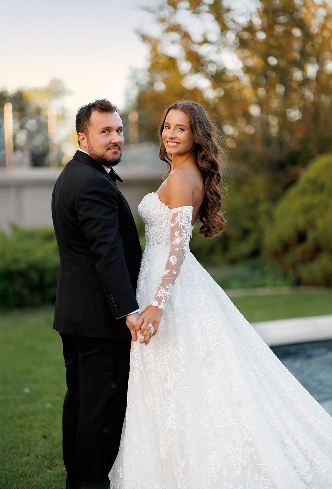 Couple in wedding attire hold hands outdoors, bride in lace gown, groom in black tuxedo, smiling.