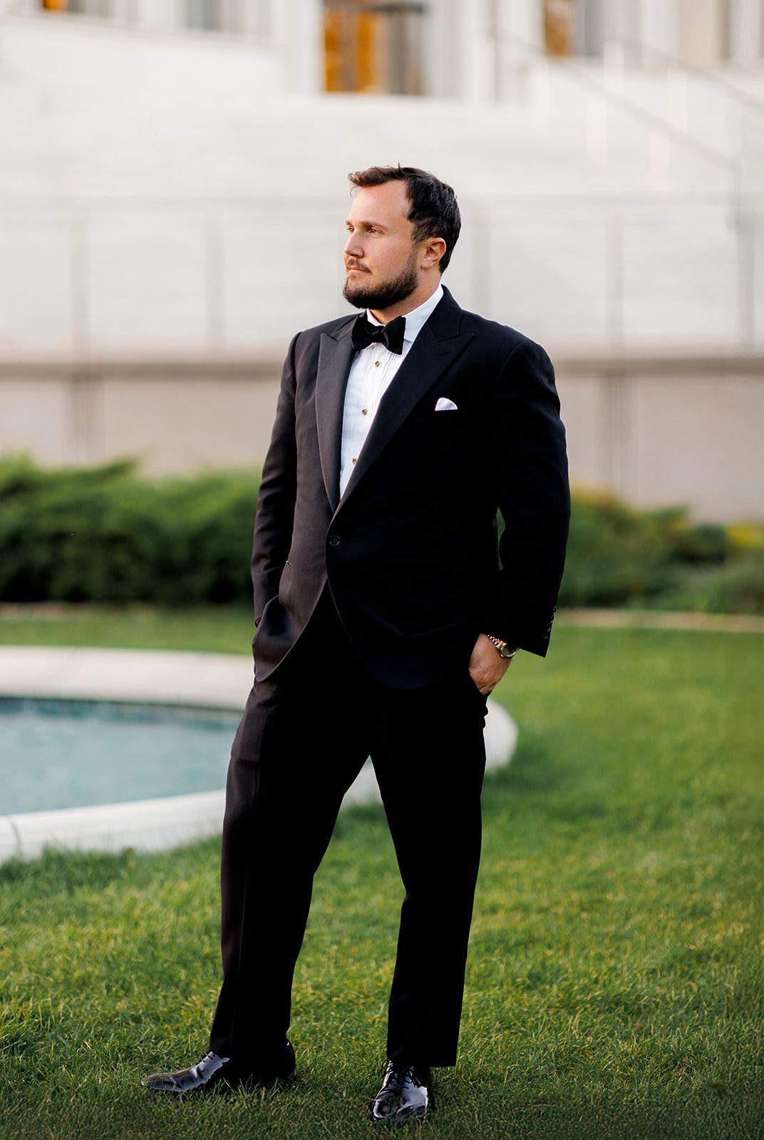 Dapper groom in a classic black tuxedo with bow tie stands confidently on a green lawn.