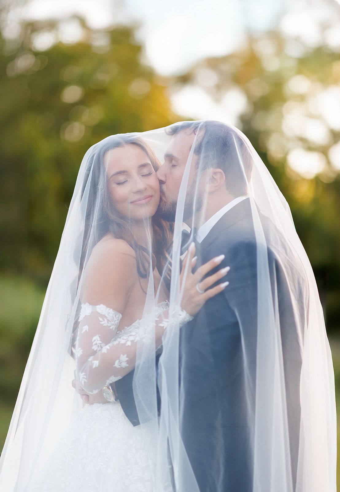 Bride and groom share a tender kiss under a flowing wedding veil outdoors during golden hour.