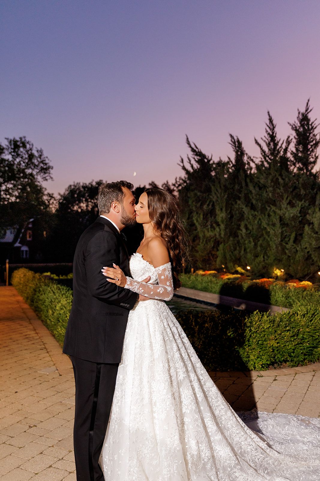 Bride and groom share a romantic kiss at twilight in a lush garden, she wearing an elegant off-shoulder lace gown.