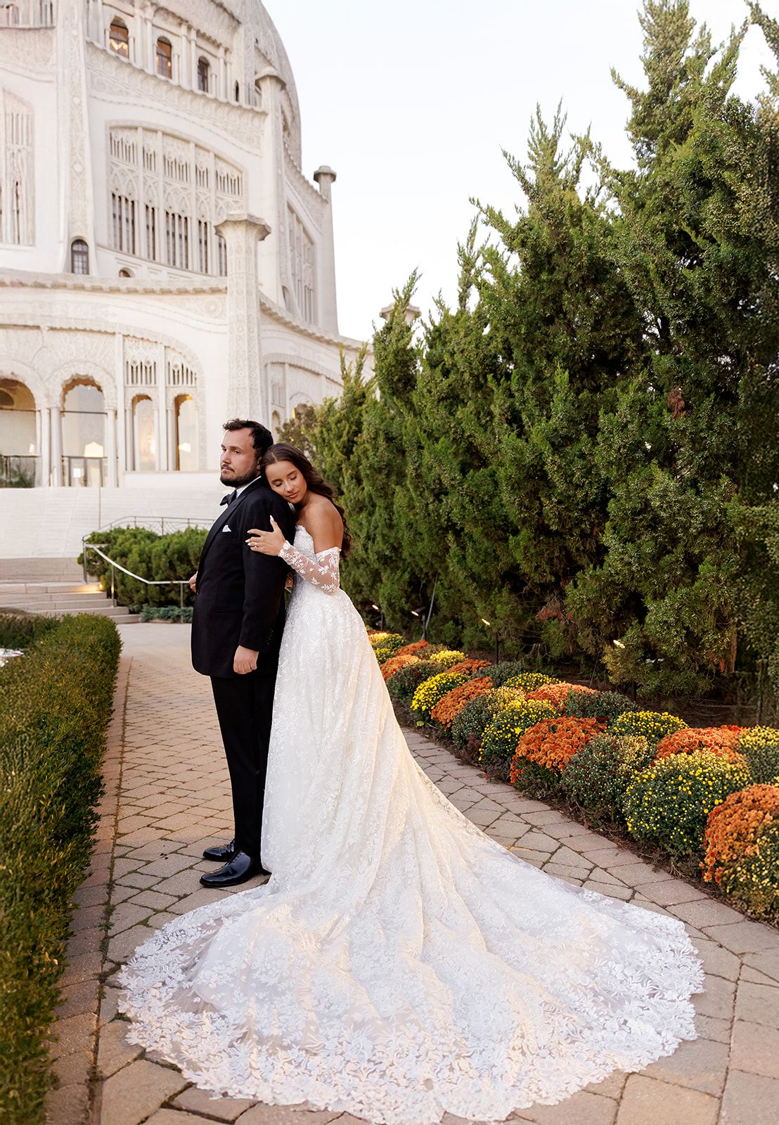 Bride in lace gown with long train poses with groom in black tux outside ornate white building with colorful gardens.