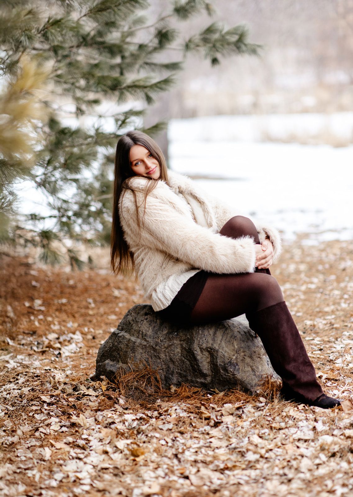 Young woman in cream fuzzy jacket and brown boots seated on stone rock in snowy autumn forest landscape.