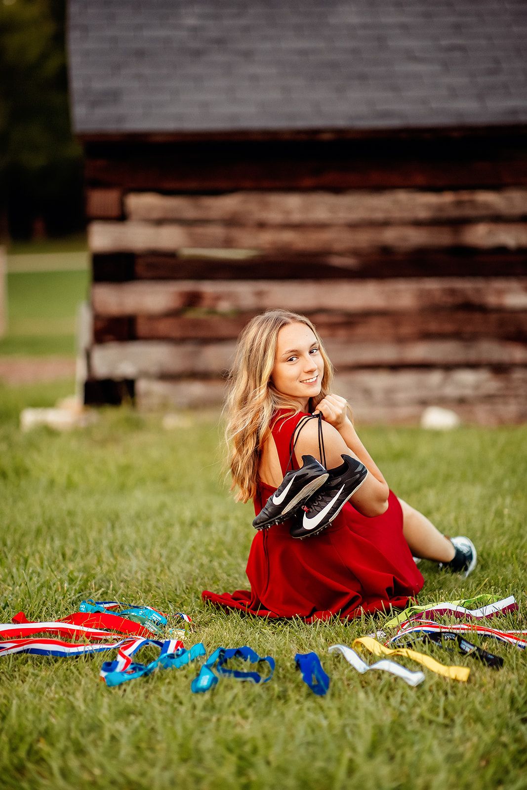 A smiling teenage girl in a red dress sits on grass holding Nike cleats, surrounded by colorful athletic medals and ribbons.