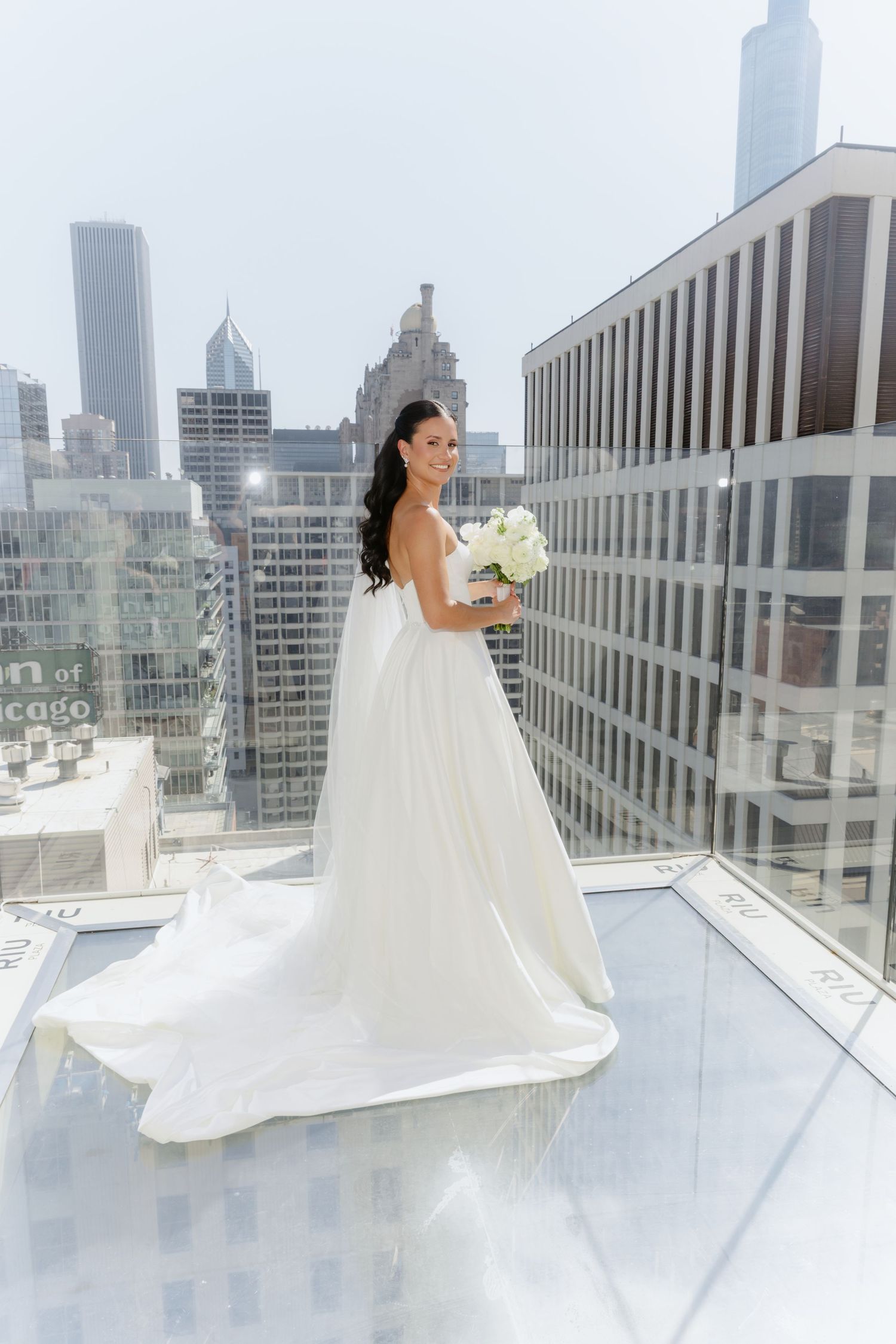 Stunning bride in flowing white ballgown holds white bouquet on rooftop with Chicago city skyline in background.