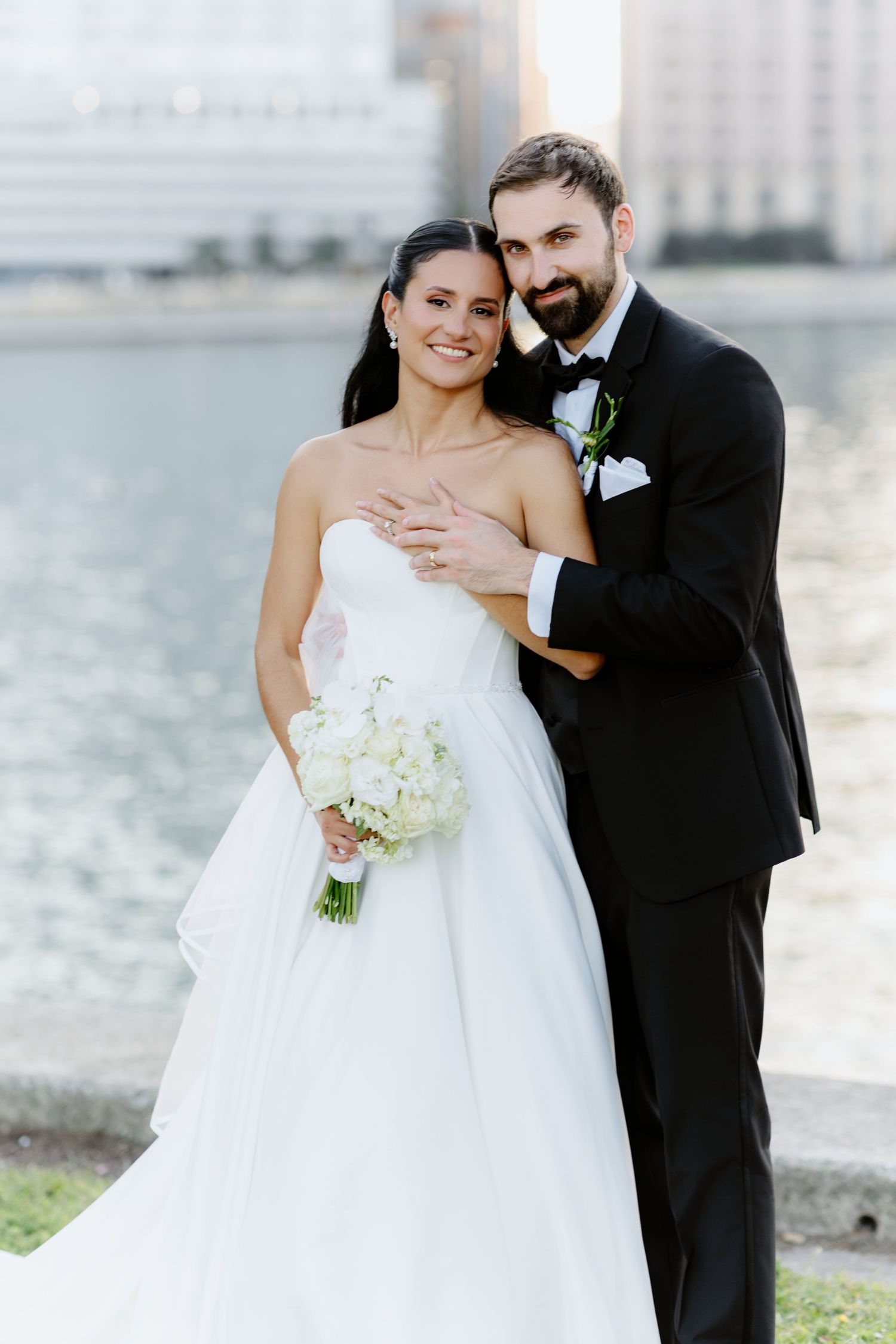 Smiling bride and groom embracing by urban waterfront, bride holding white bouquet.