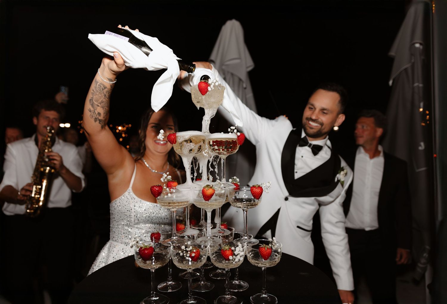Bride and groom pouring champagne tower with strawberries at outdoor wedding reception.