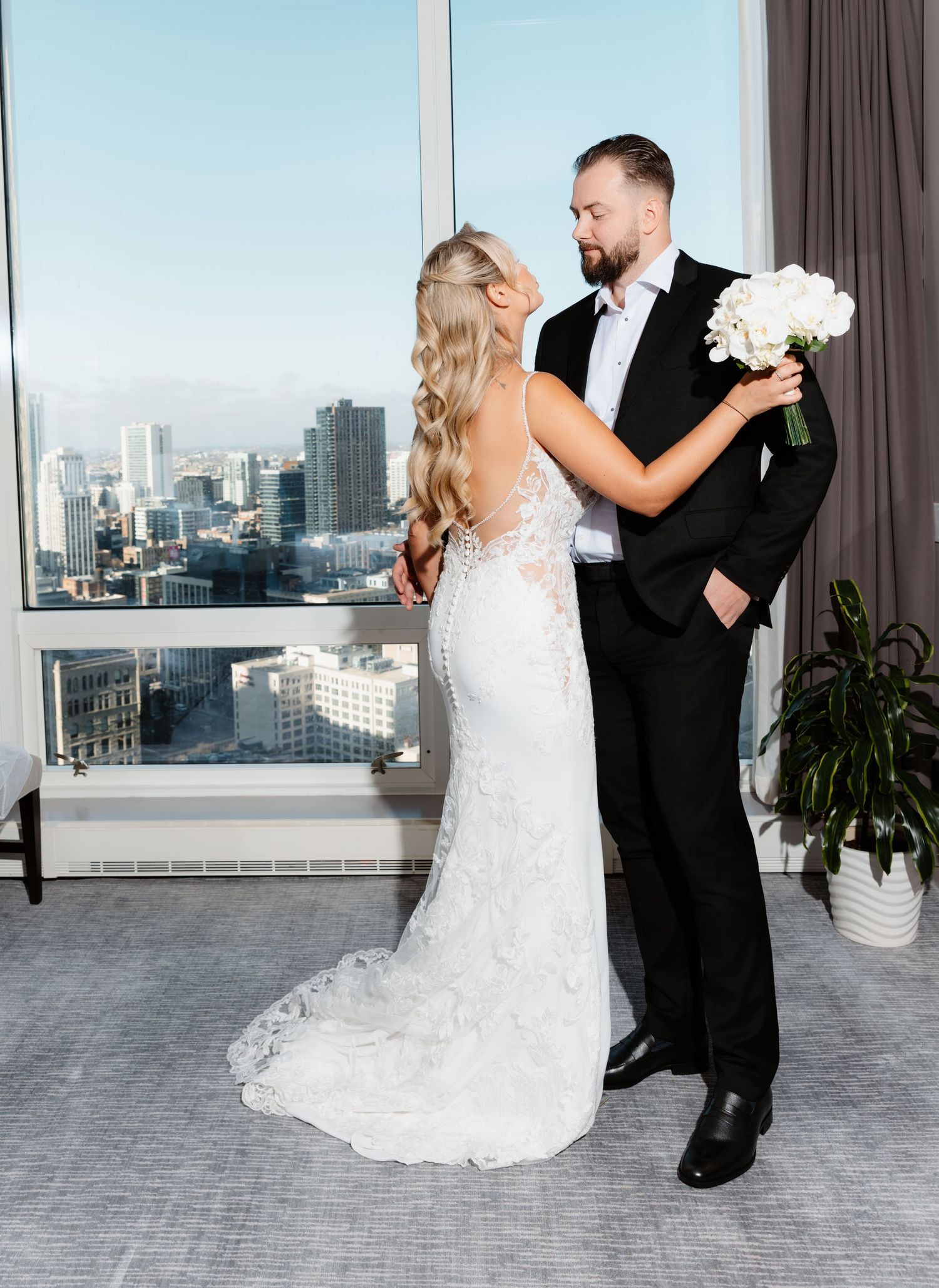 Bride and groom embrace tenderly with city skyline visible through large hotel room windows.