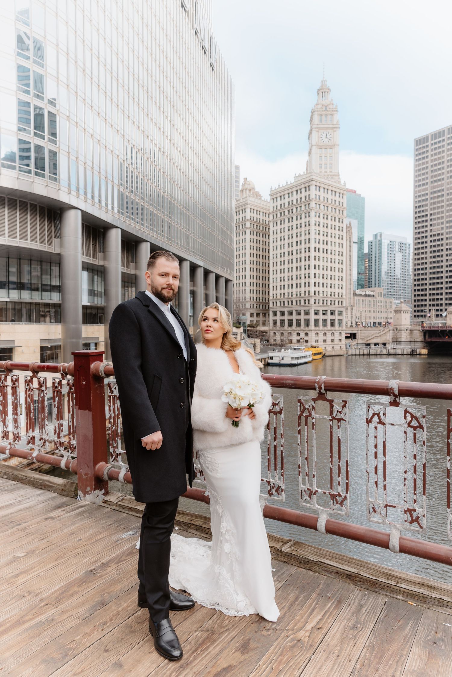 Wedding couple poses on Chicago riverwalk boardwalk with Wrigley Building and river in background.