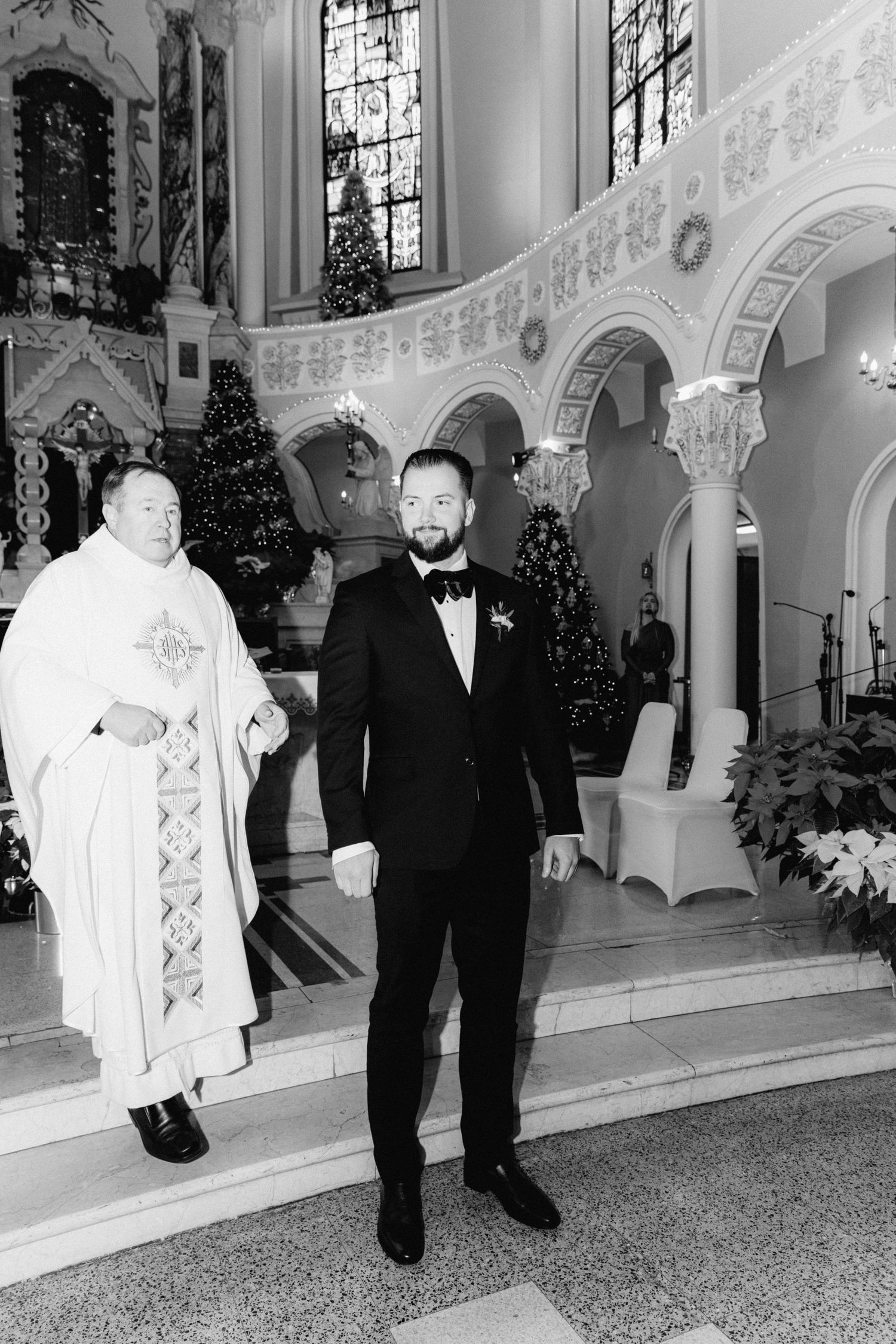 A groom in a black tuxedo stands at the altar with a priest in white vestments in a decorated church.