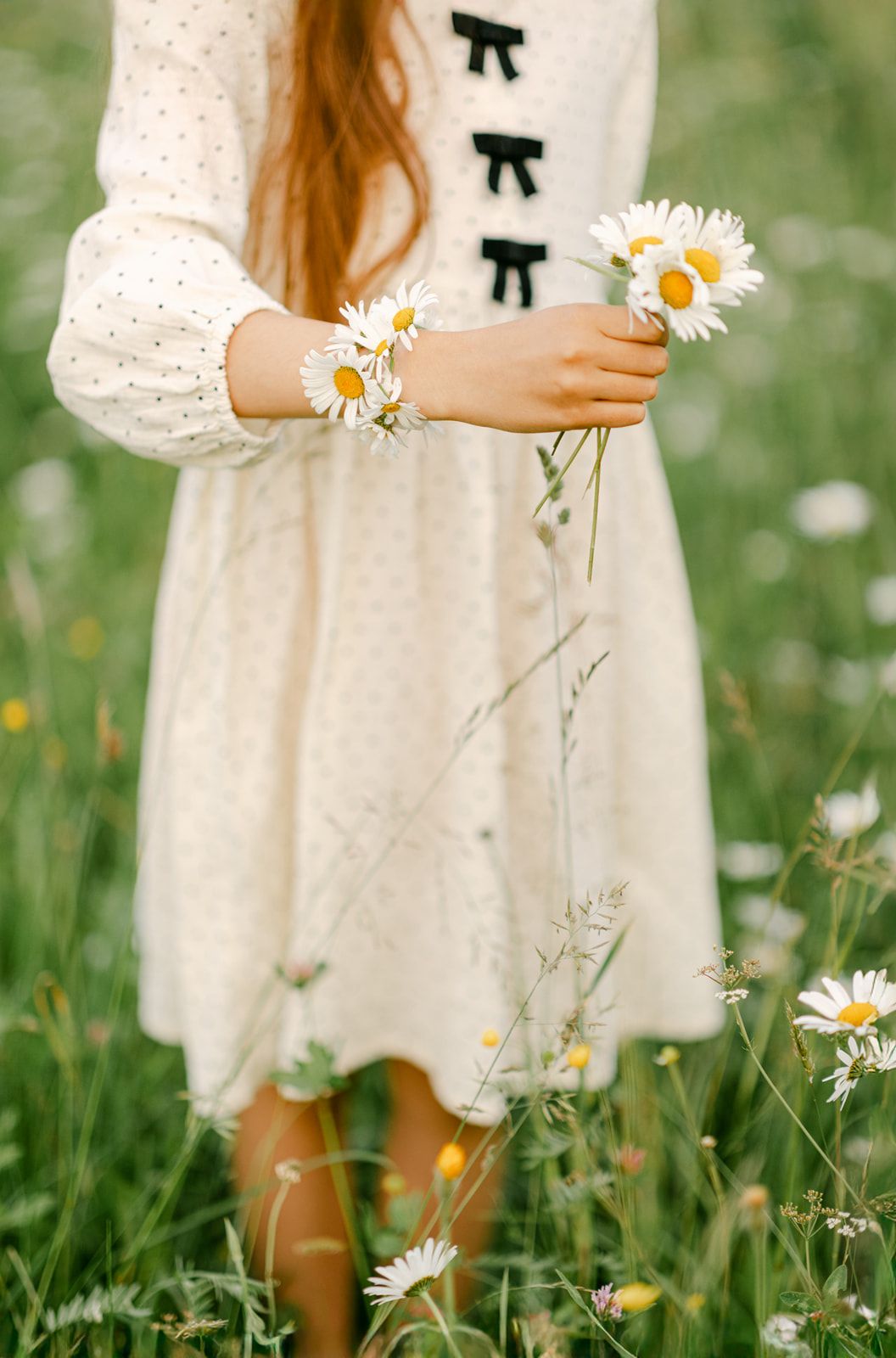 A young woman in a white polka dot dress holds fresh daisies in a lush green meadow filled with wildflowers.