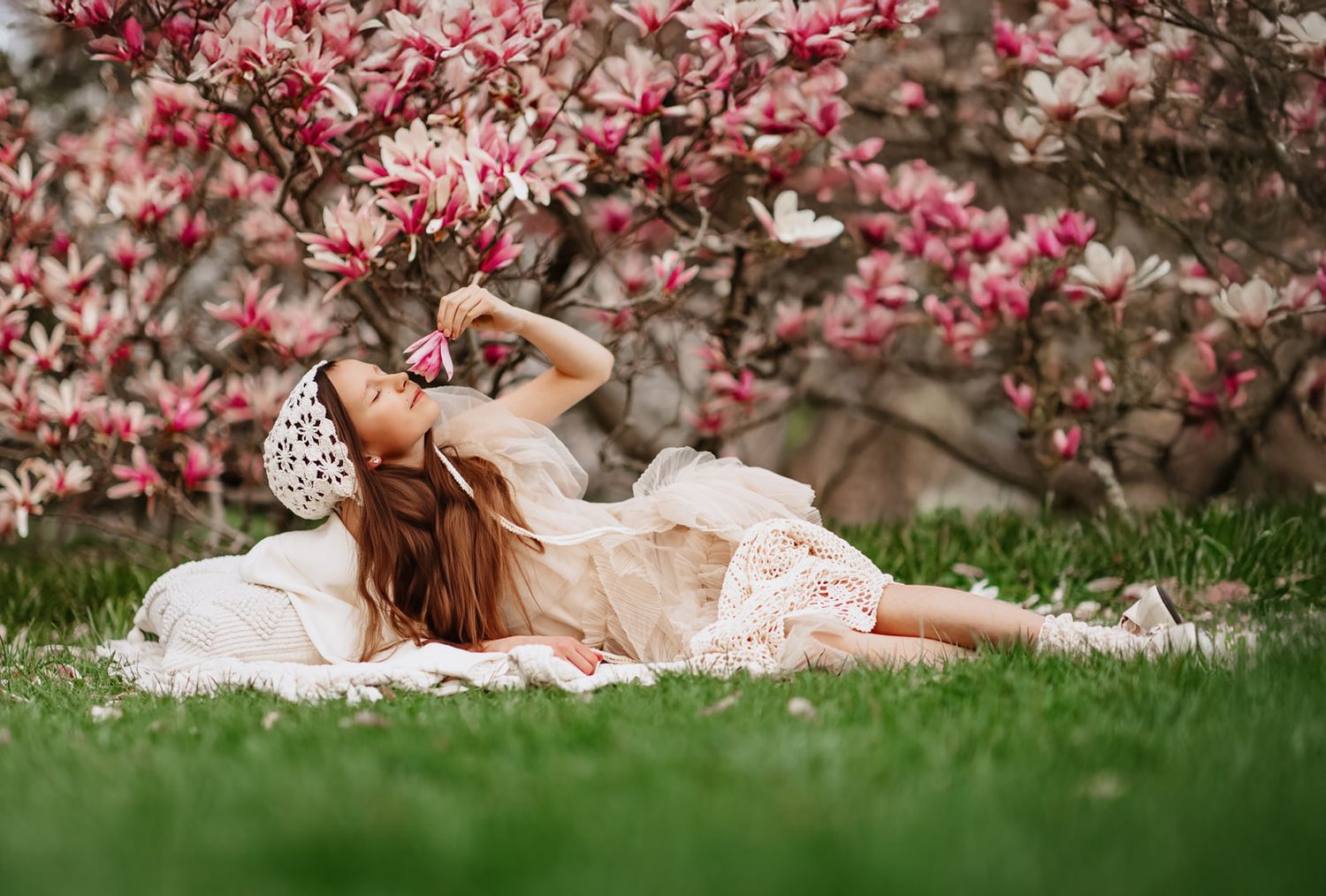 Young woman in white lace dress lying on grass beneath blooming pink magnolia tree in spring garden.