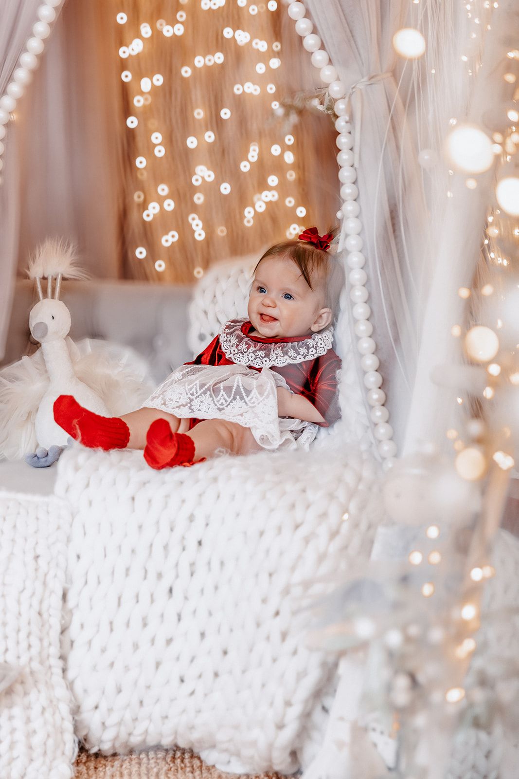 Baby girl in red and white dress sits on white knit blanket under glowing fairy light canopy with stuffed animal toy.