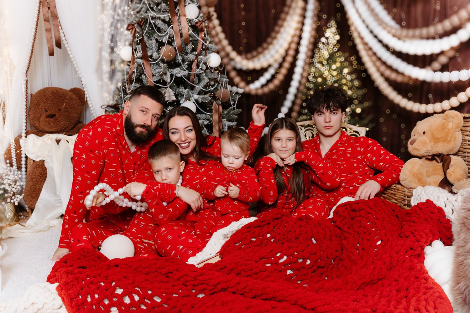 Family of five in matching red Christmas pajamas sitting on a bed decorated with a festive tree and holiday decor.
