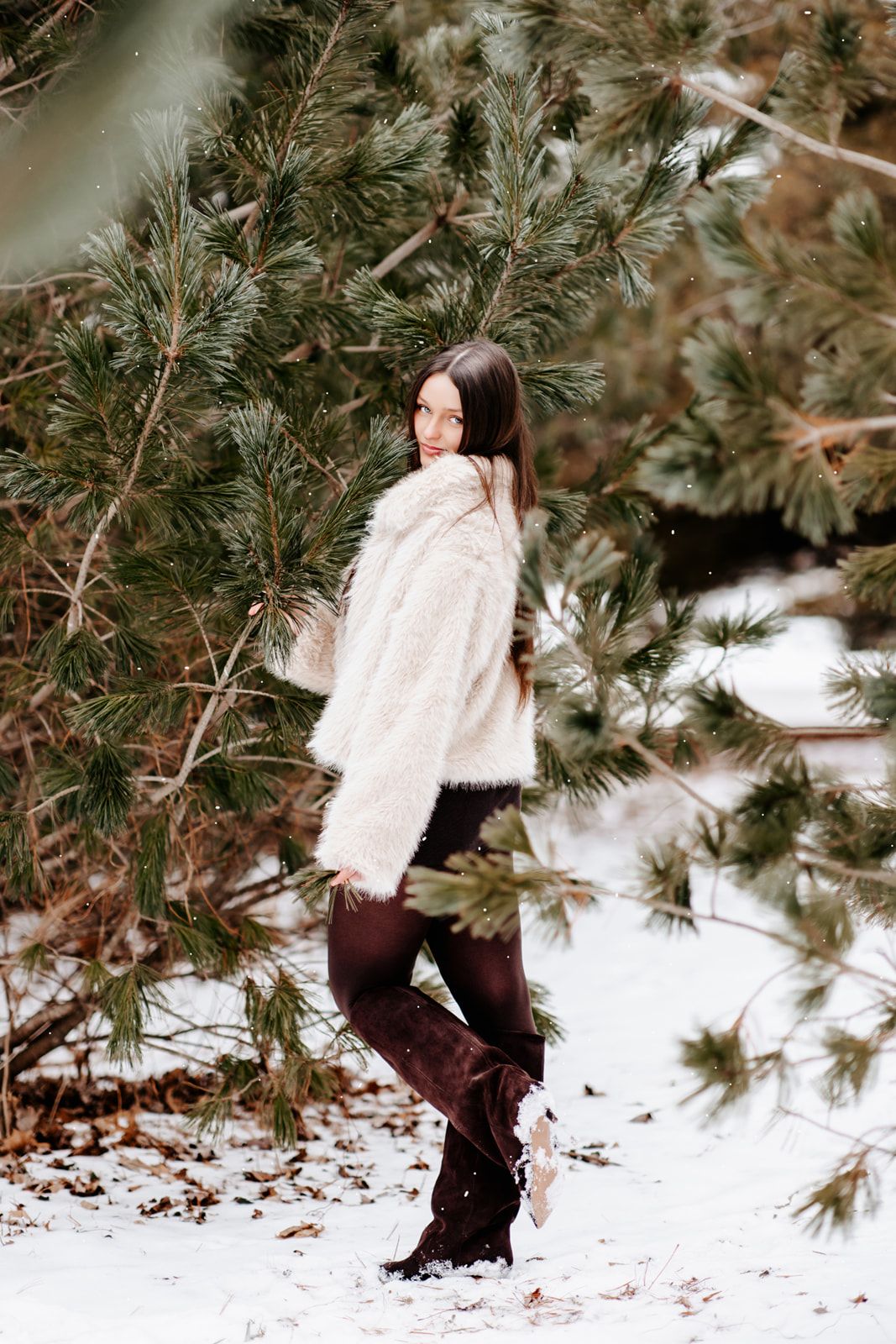A young woman in a white faux fur coat and dark boots stands among snow-covered pine tree branches in winter.