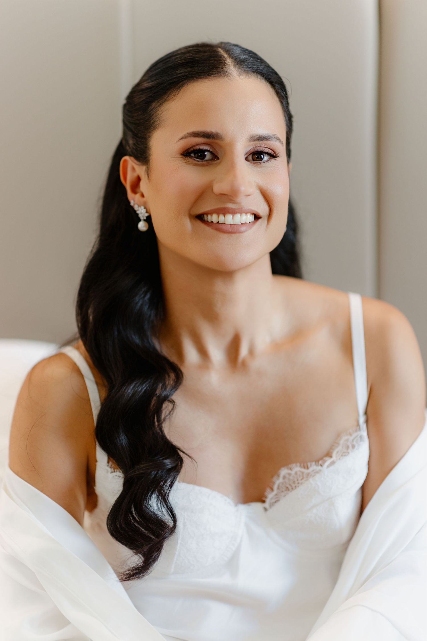 Smiling bride with dark wavy hair, pearl earrings, and white lace bridal lingerie on her wedding day.