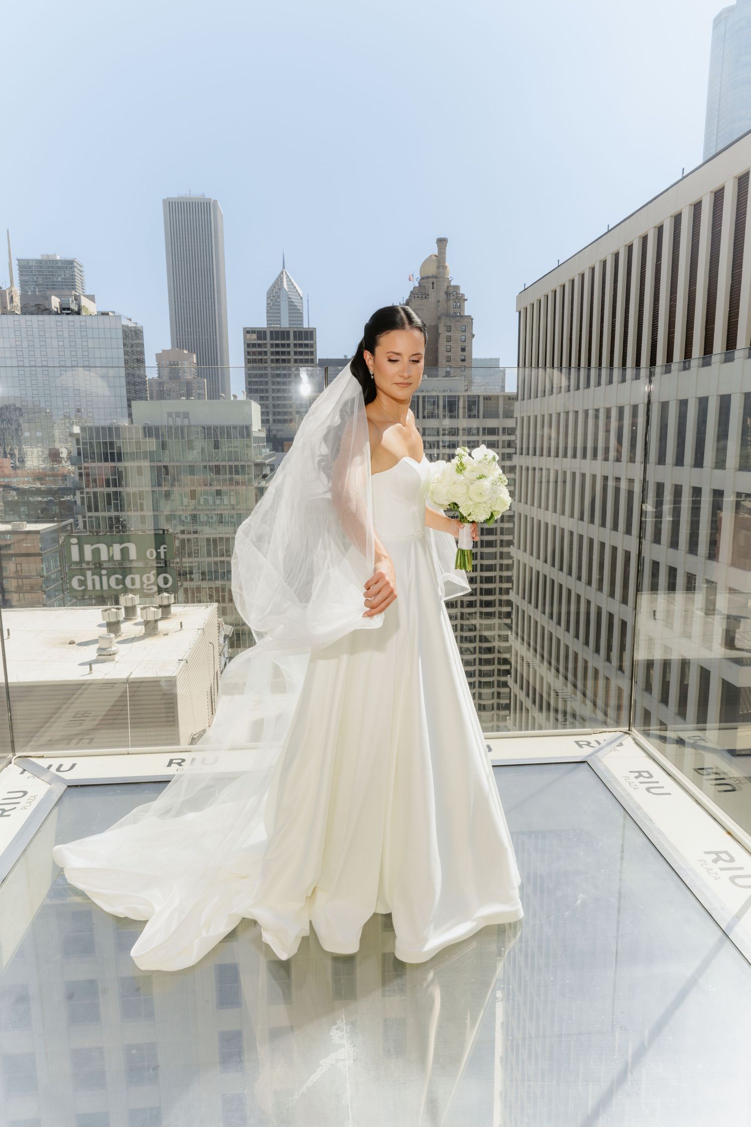 Elegant bride in white gown and veil holding bouquet on rooftop with Chicago skyline.
