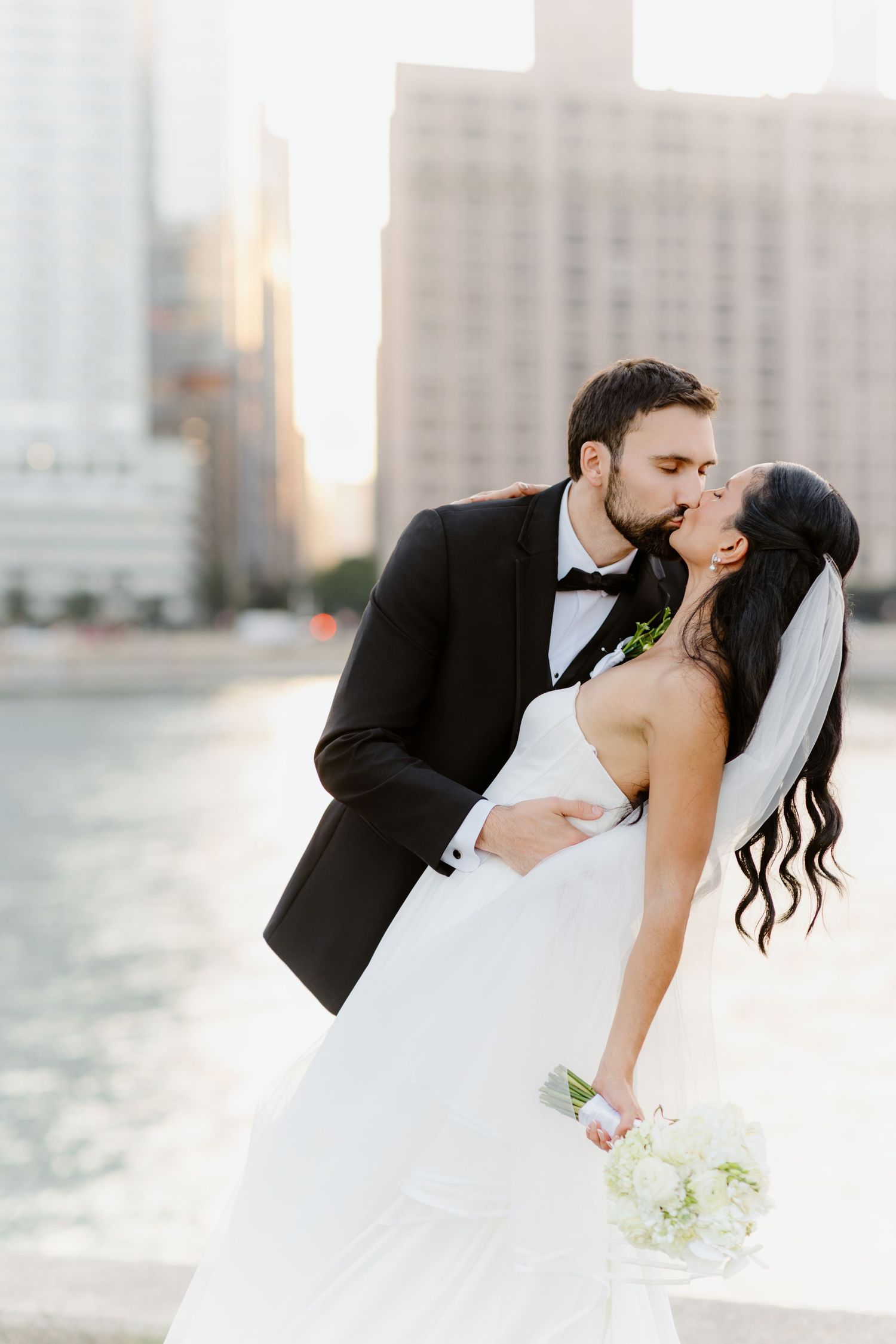 Groom dips bride for a romantic kiss by the river with sunlit city skyscrapers in the background.