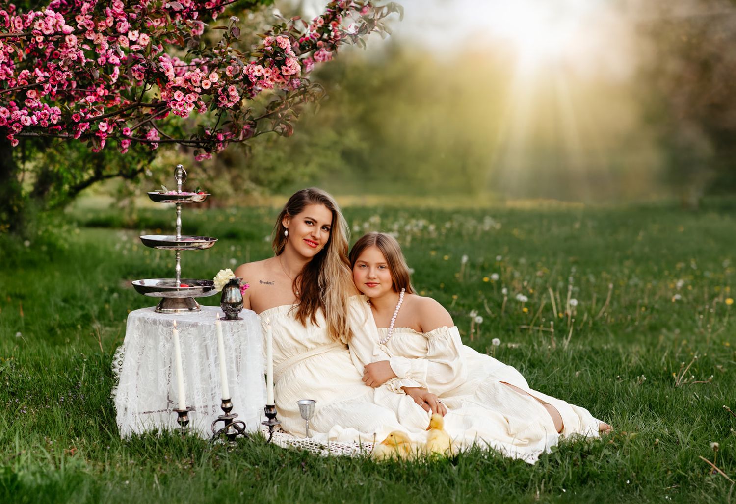 Woman and girl in white dresses sit on grass during outdoor picnic under pink blossoming tree in golden sunlight.