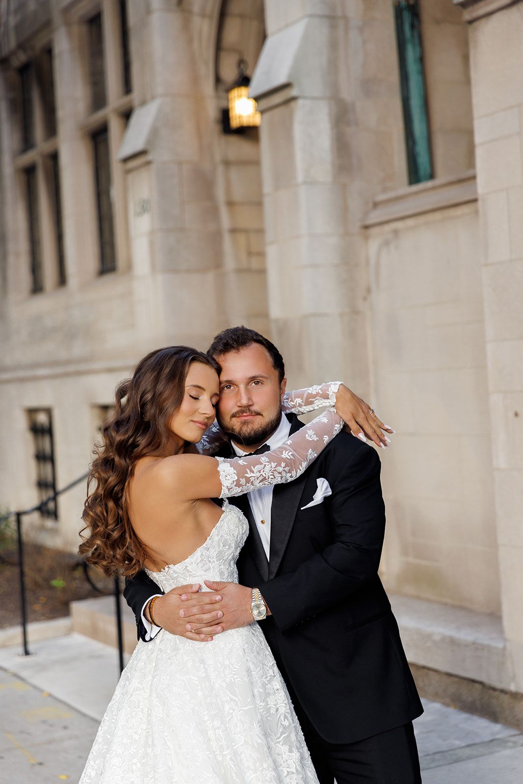 Bride in backless lace gown embraces groom in black tuxedo outside historic stone building.