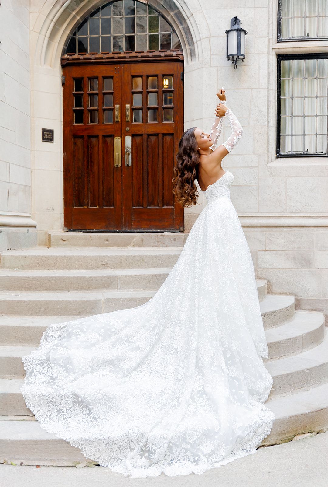 Bride in lace wedding gown with long train poses on stone steps before ornate wooden doors.