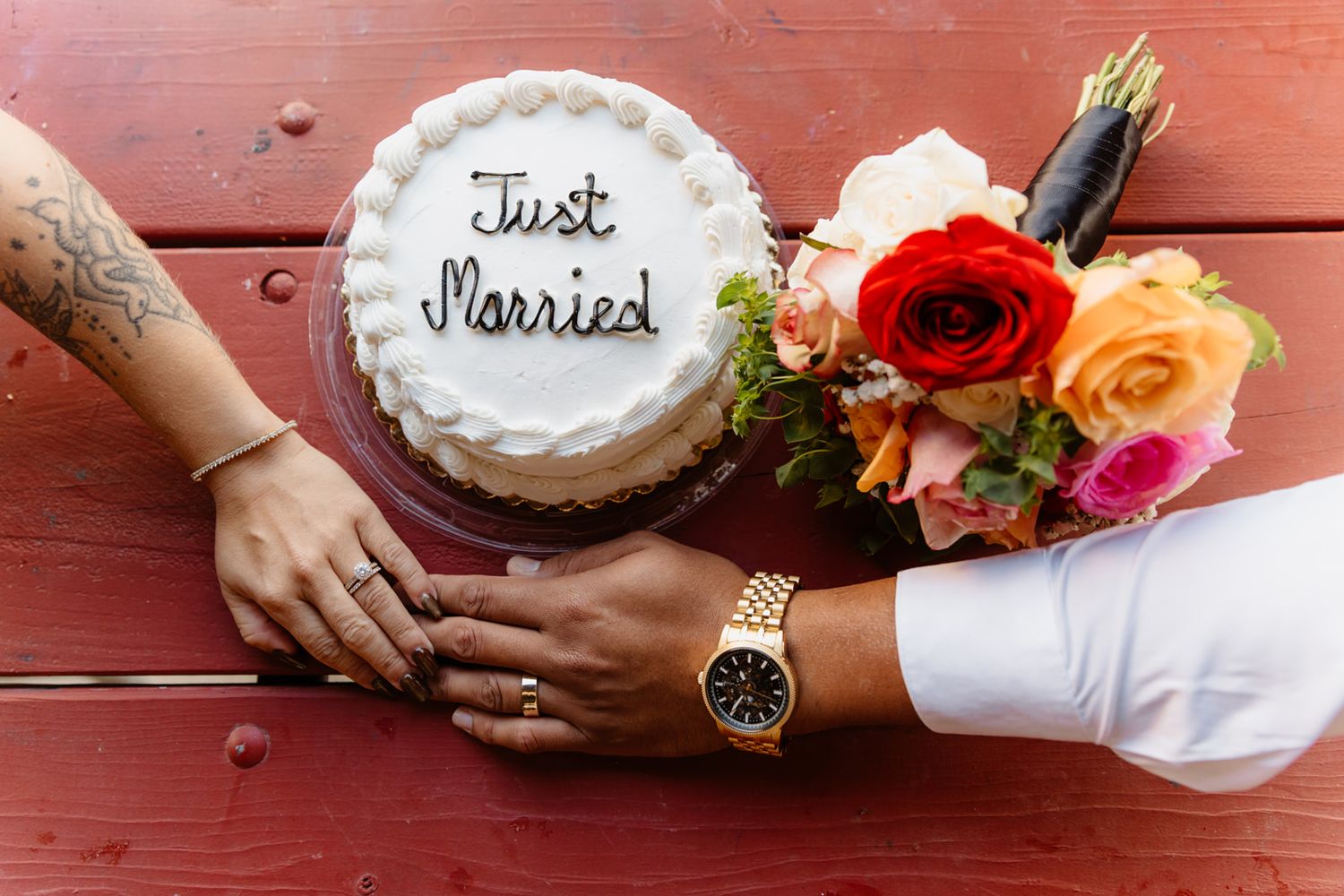 Newlyweds hands beside a Just Married white wedding cake and colorful bridal bouquet on a red wooden table.
