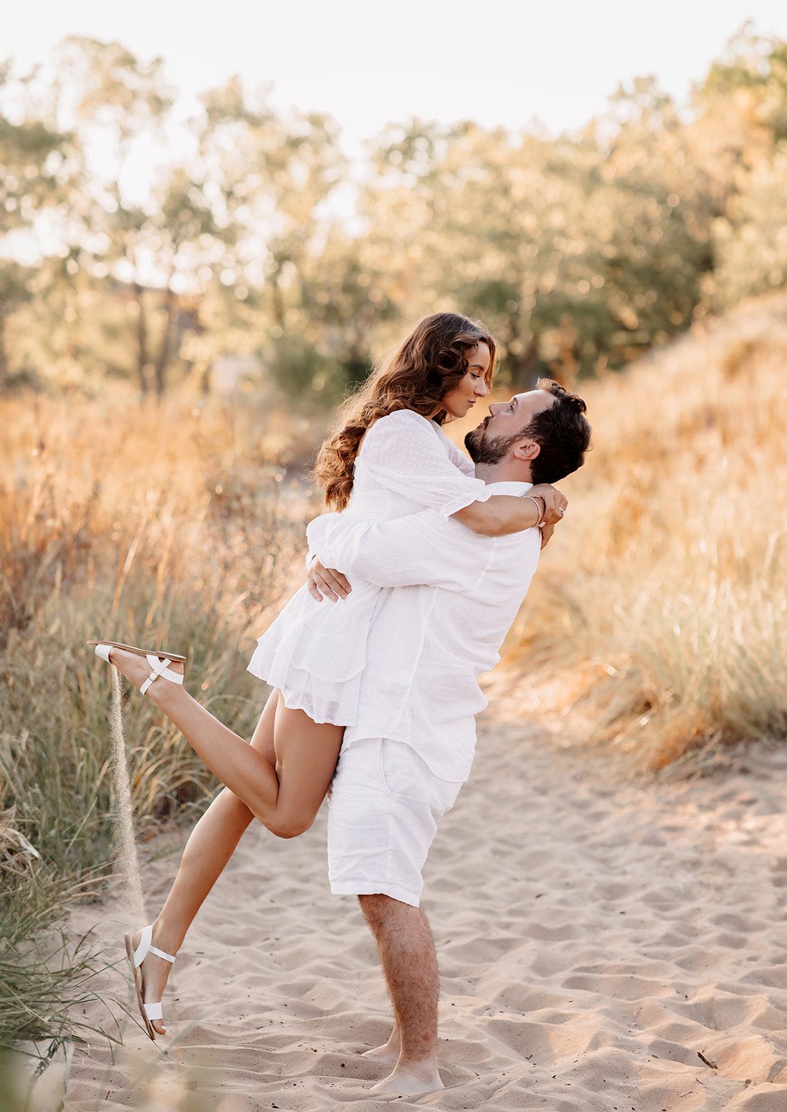 Man lifting woman in white dress on sandy beach path surrounded by golden autumn foliage.