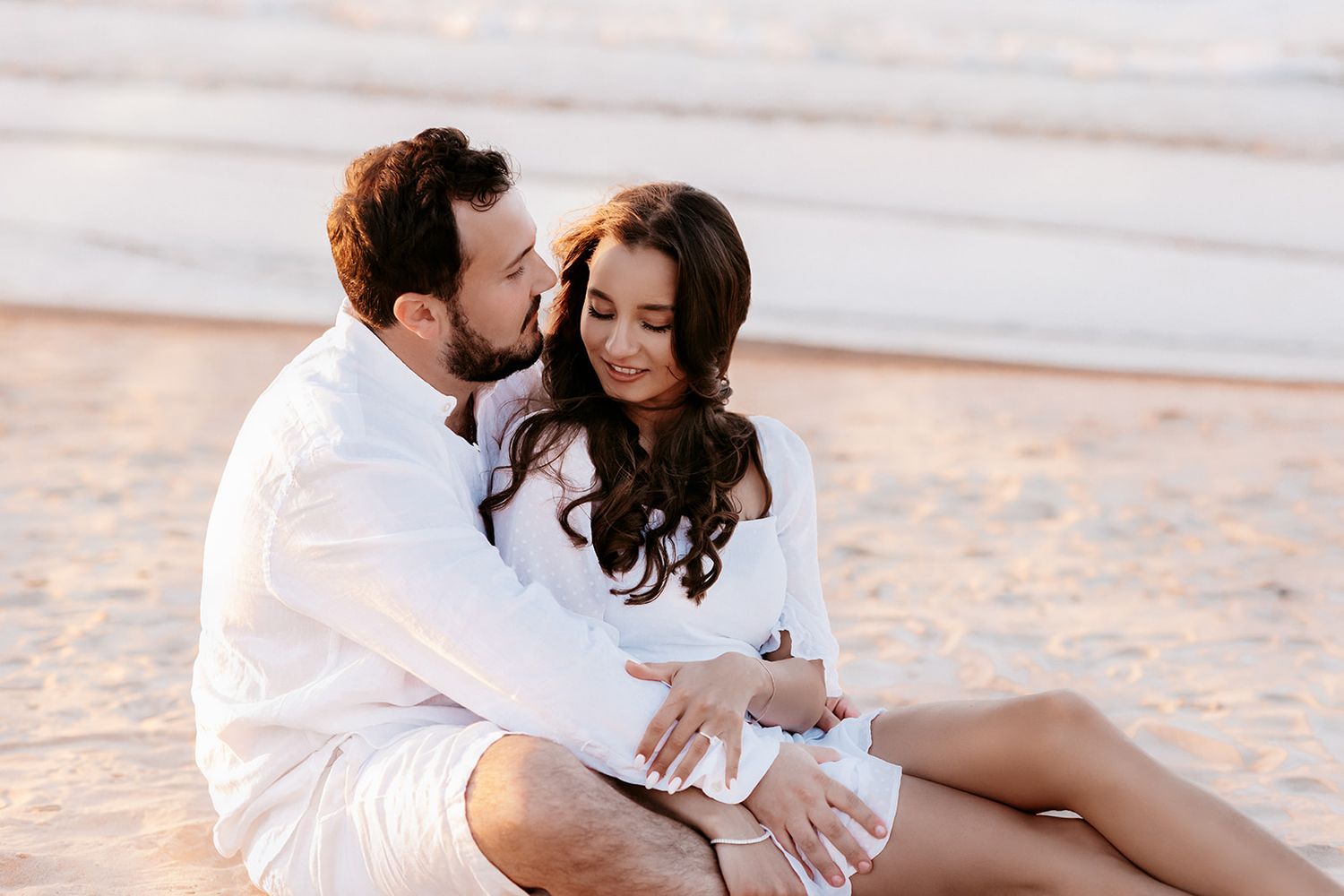 Couple sitting on golden sand beach at sunset, sharing a tender romantic moment.