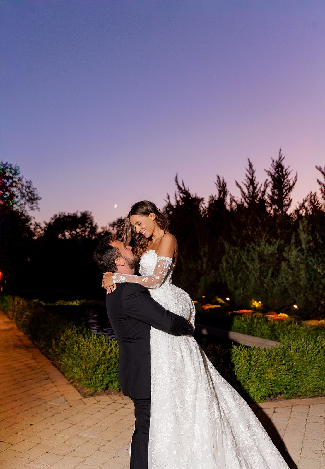 Groom lifts bride in white lace gown at dusk, sharing romantic moment against purple twilight sky.