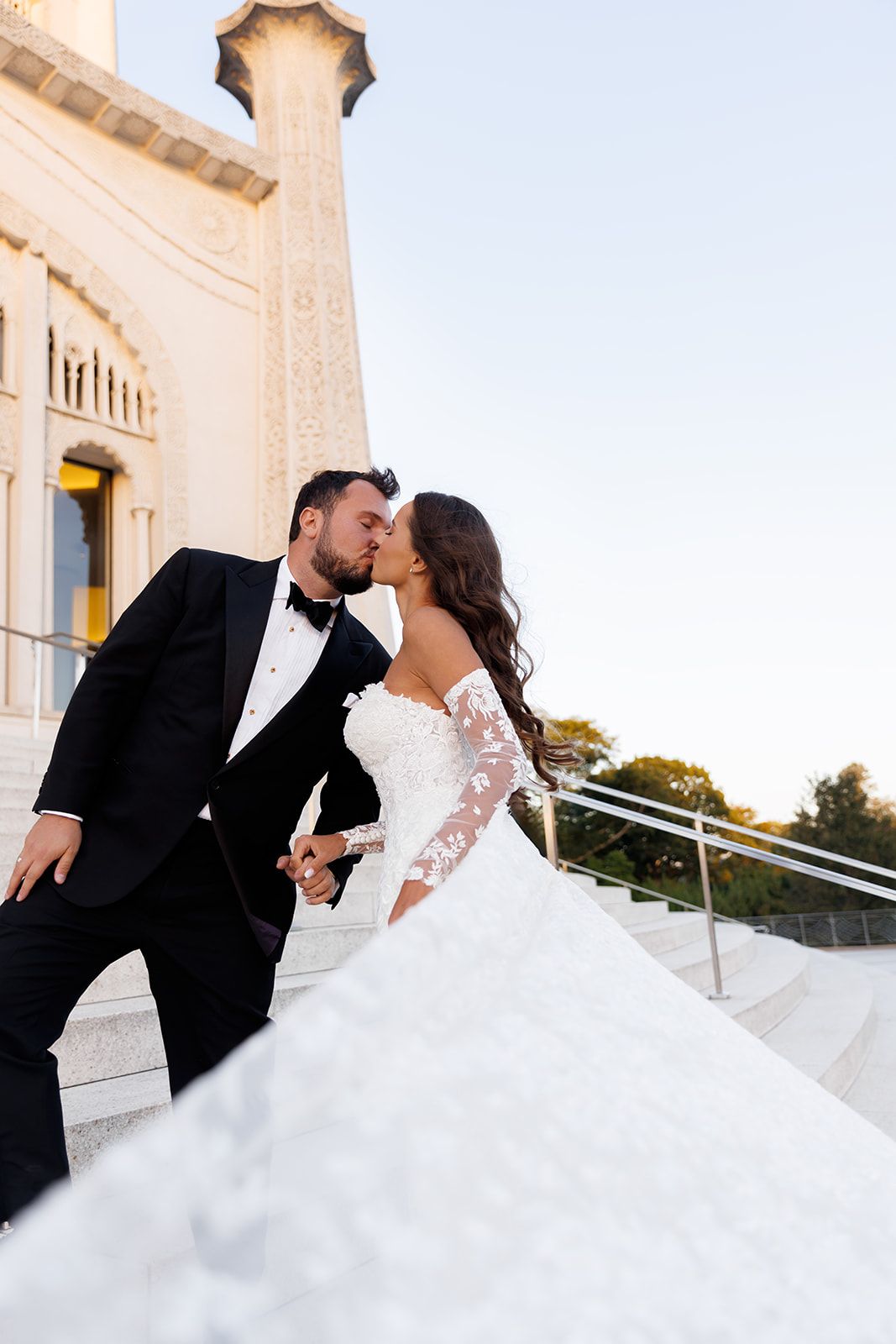 Bride and groom share a romantic kiss on outdoor steps, bride in lace off-shoulder gown, groom in black tuxedo.