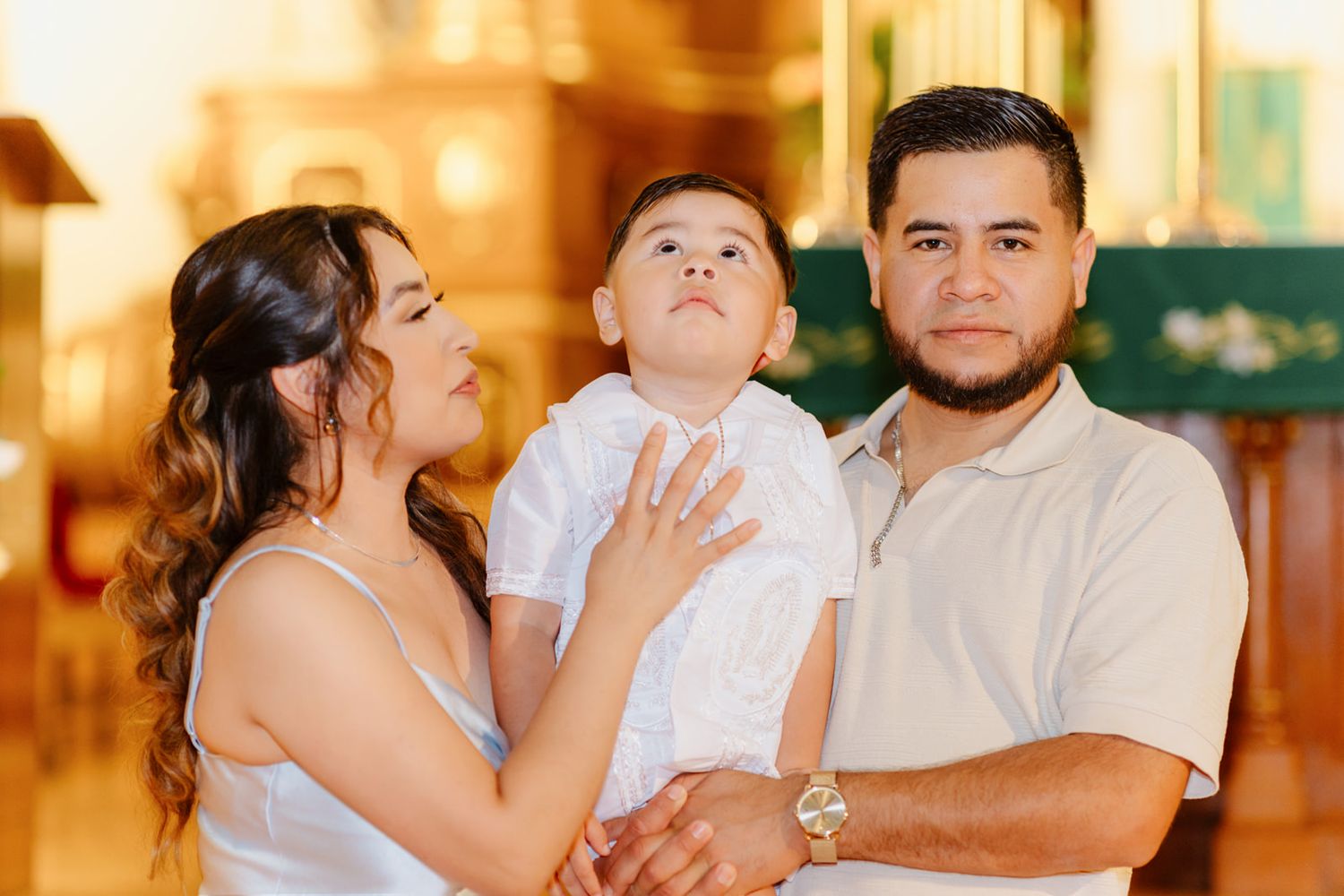 A smiling family with a baby boy in white baptism attire poses inside a warmly lit church.