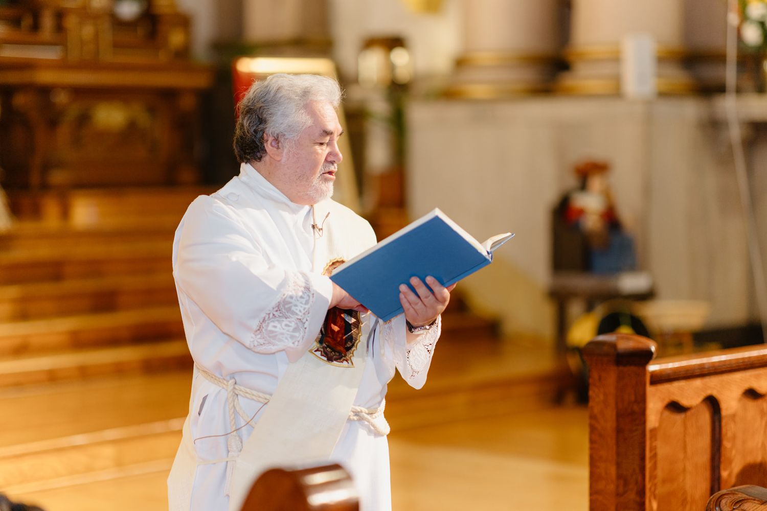 Elderly priest in white robes reads aloud from a blue liturgical book inside a Catholic church.