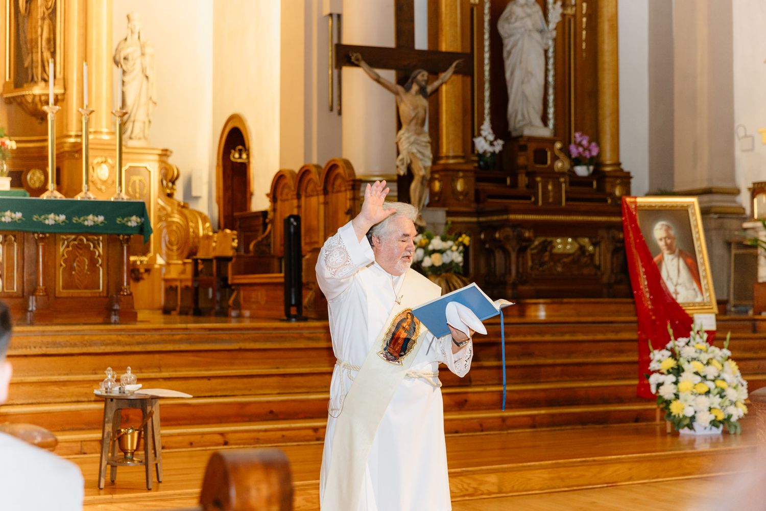 Catholic priest in white robes raises hand while holding book during church ceremony with altar and crucifix in background.