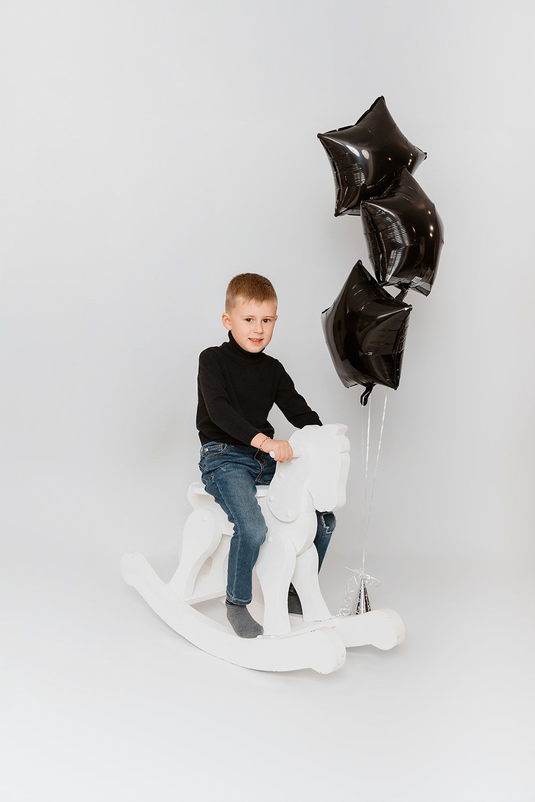 Young boy in black sweater and jeans sitting on white rocking horse with black star-shaped balloons on white background.