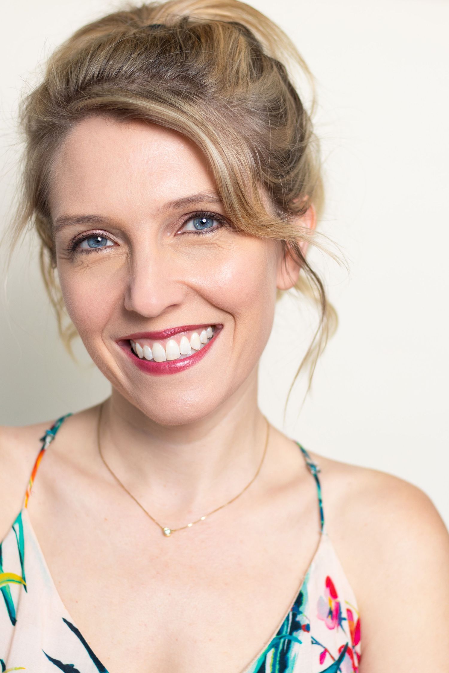 Happy blonde woman with blue eyes and updo smiling brightly in floral dress against white background.