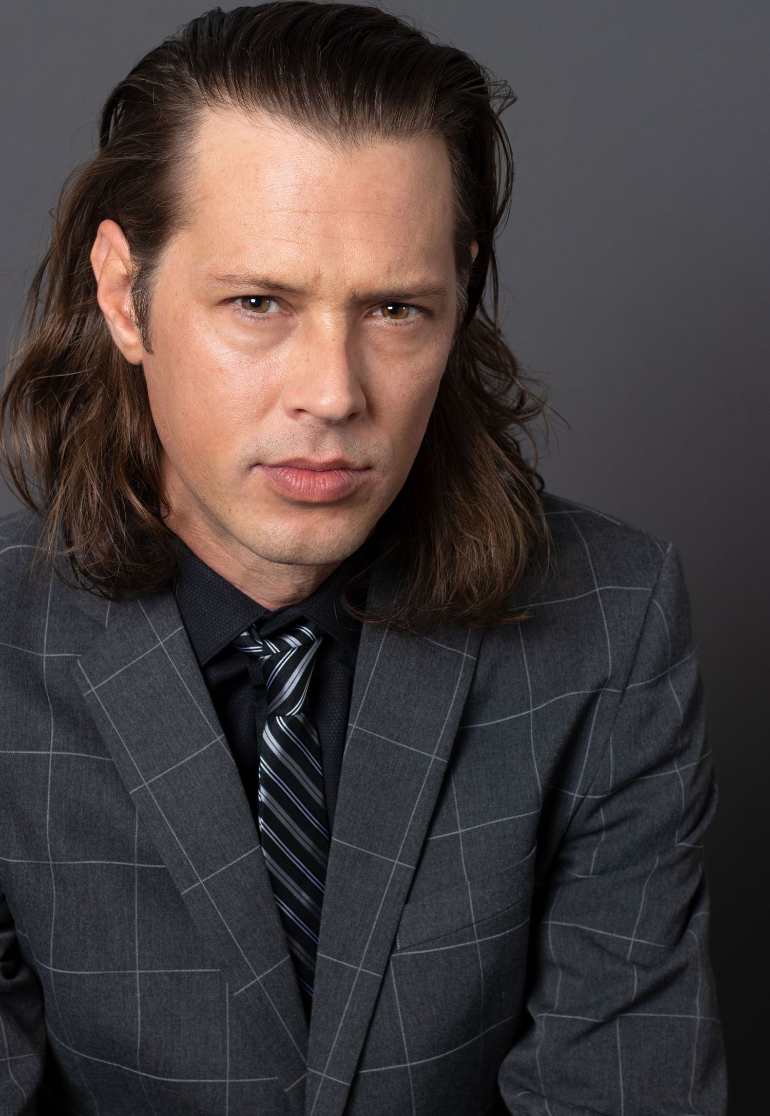 Man with long dark hair wearing a gray plaid suit and tie poses for a serious professional headshot in Charleston, SC.