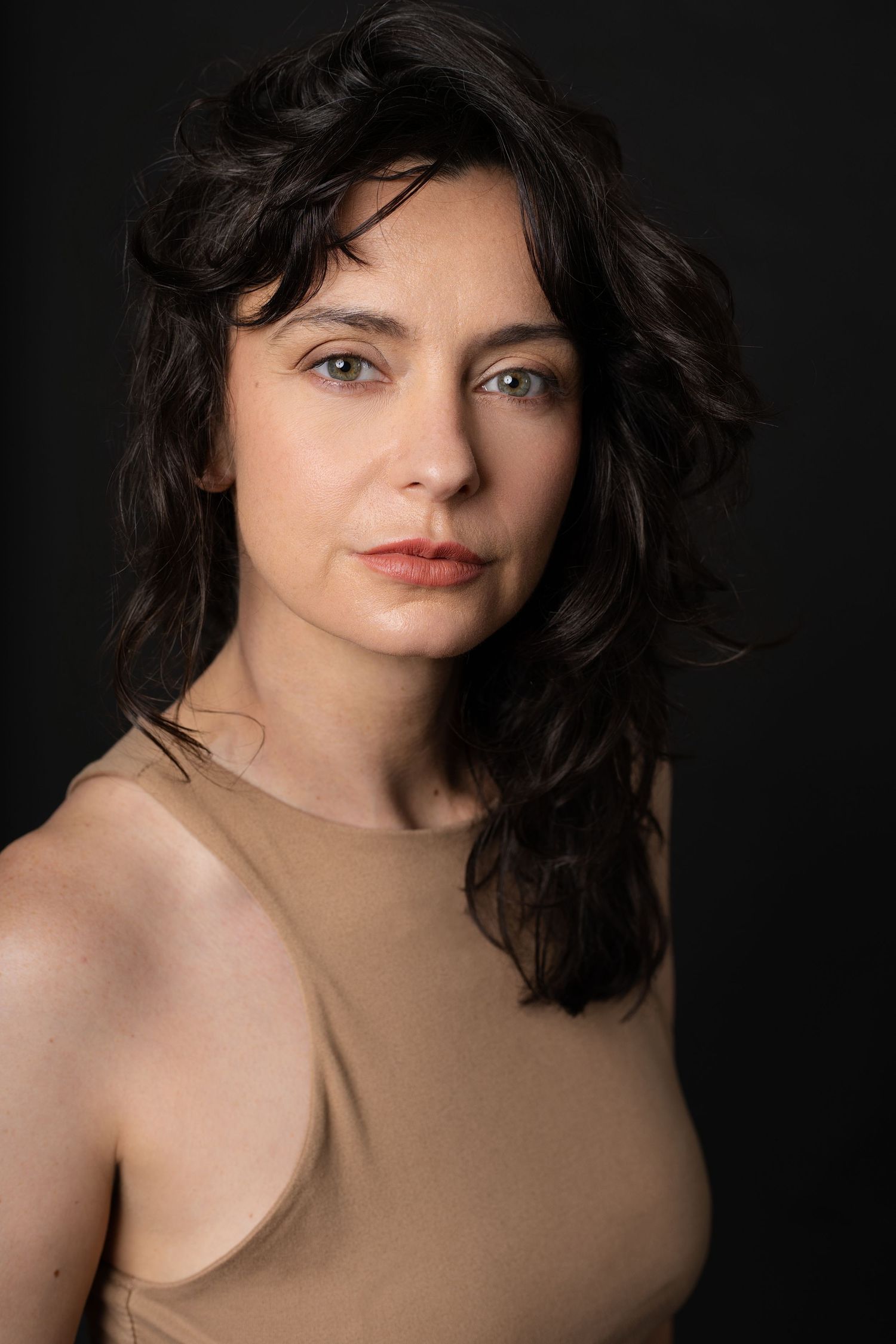 Dark-haired woman in beige tank top poses for professional actor headshot against black background in Charleston, SC.