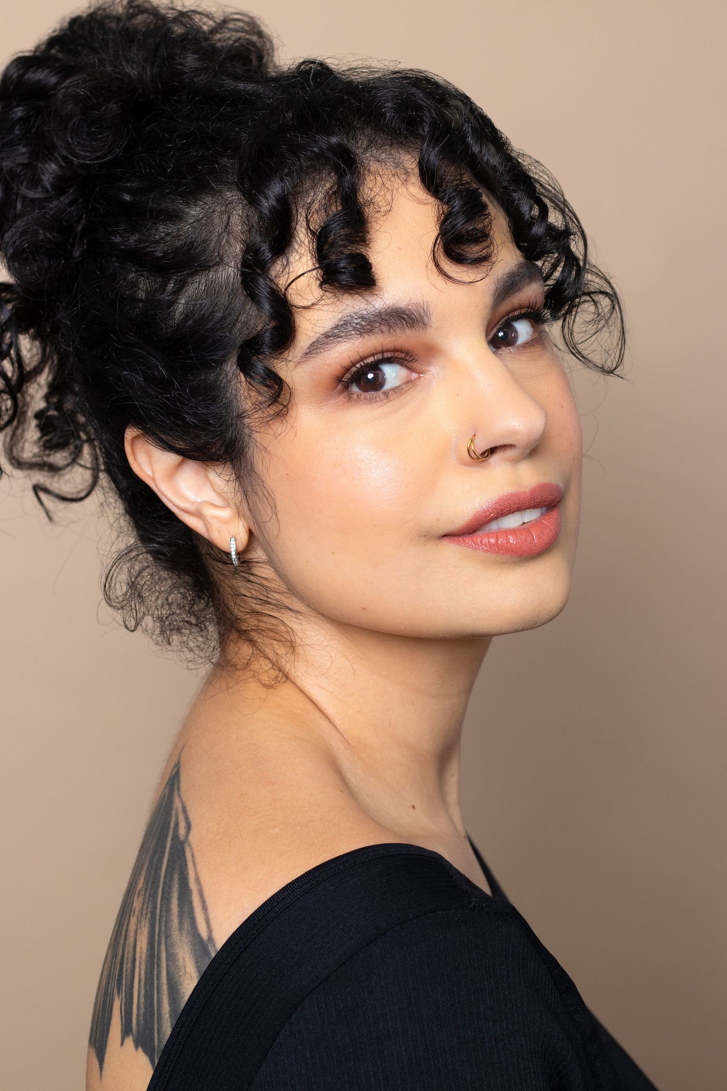 Young woman with curly black updo and nose ring showing shoulder tattoo in a professional portrait.