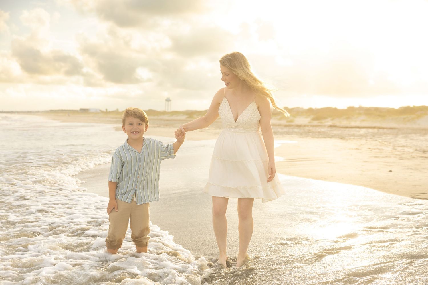Mother and young son holding hands walking through ocean waves at sunset on a sandy beach.