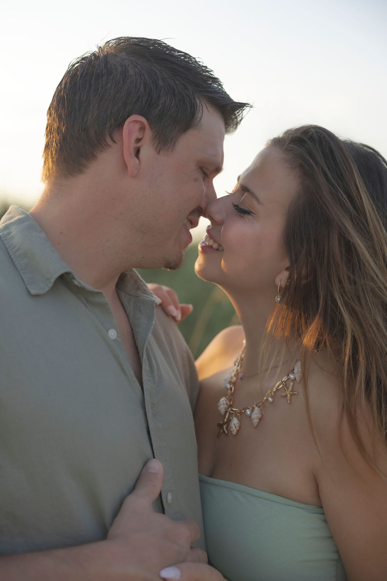 Romantic couple sharing a tender kiss outdoors at golden hour, woman wearing a shell necklace and strapless top.