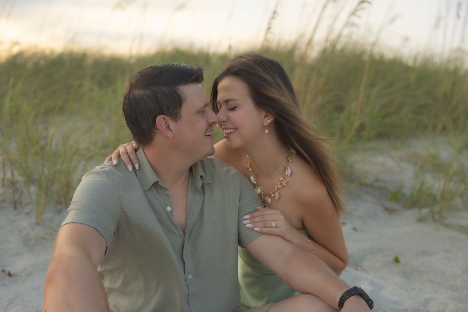 Engaged couple sitting in sand dunes at sunset, smiling at each other with engagement ring visible.