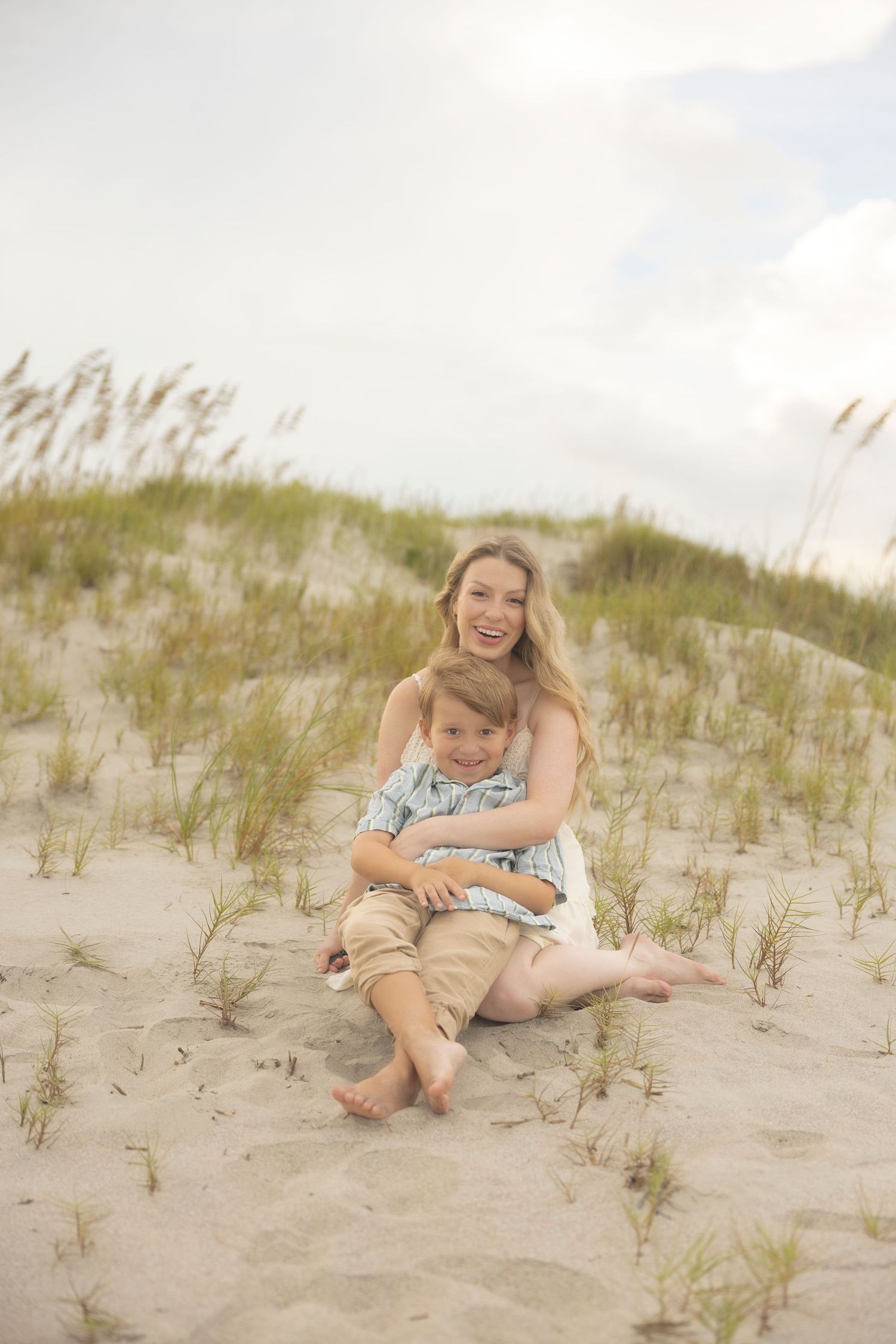 A smiling woman sits on sandy beach dunes hugging a young boy, with sea oats and cloudy sky behind them.
