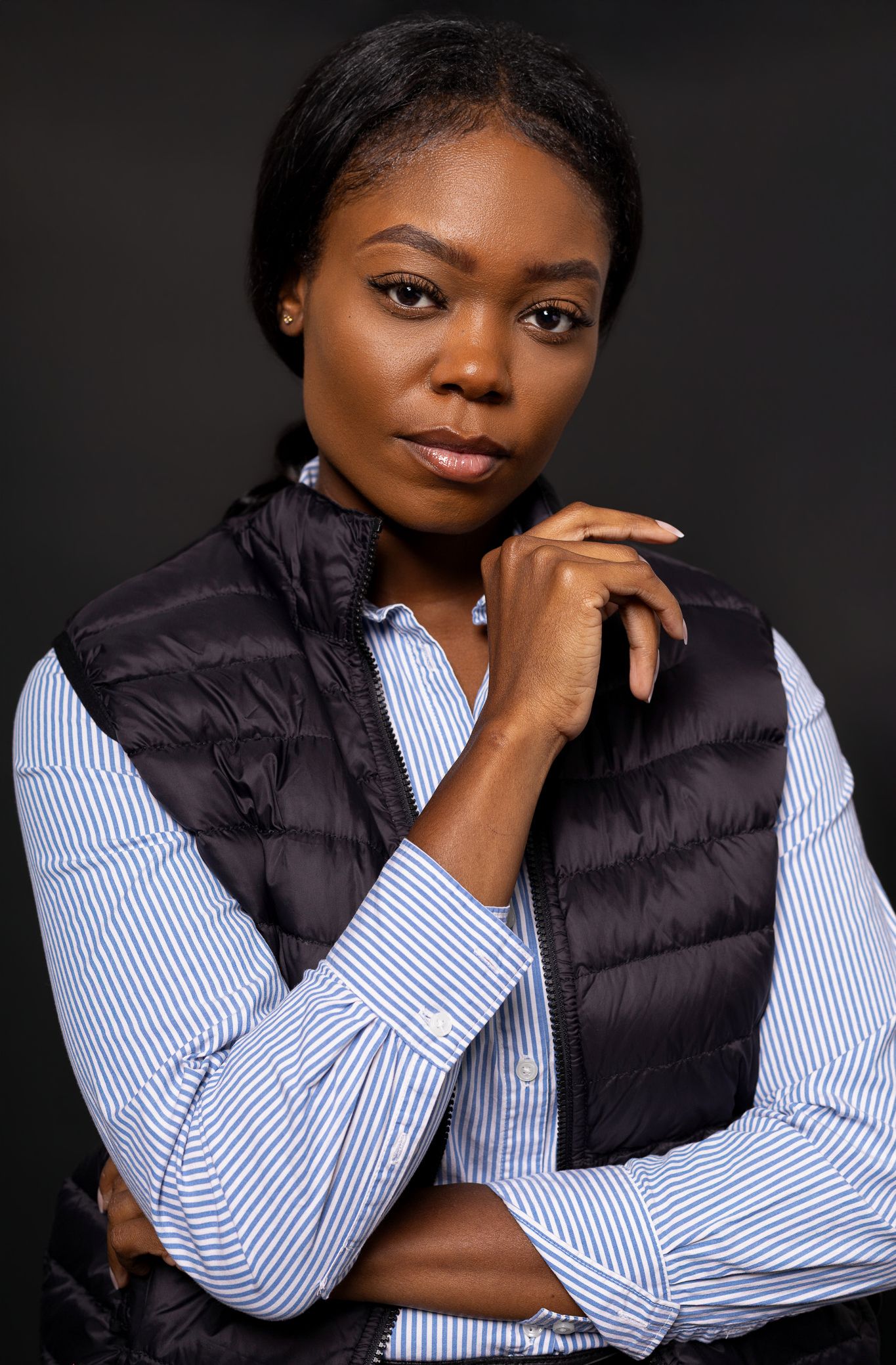 A young Black woman in a striped shirt and black puffer vest poses confidently against a dark background.