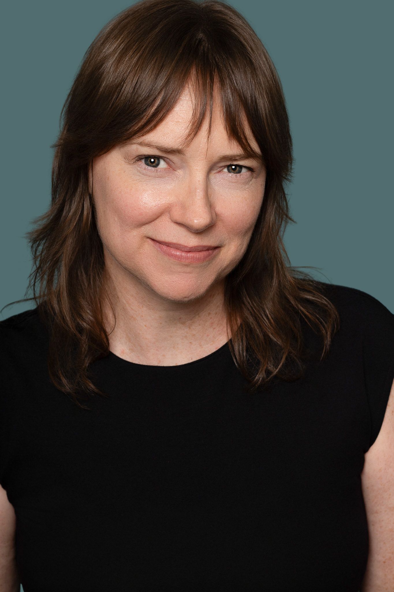 A woman with brown hair wearing a black sleeveless top poses for a professional headshot against a teal background.