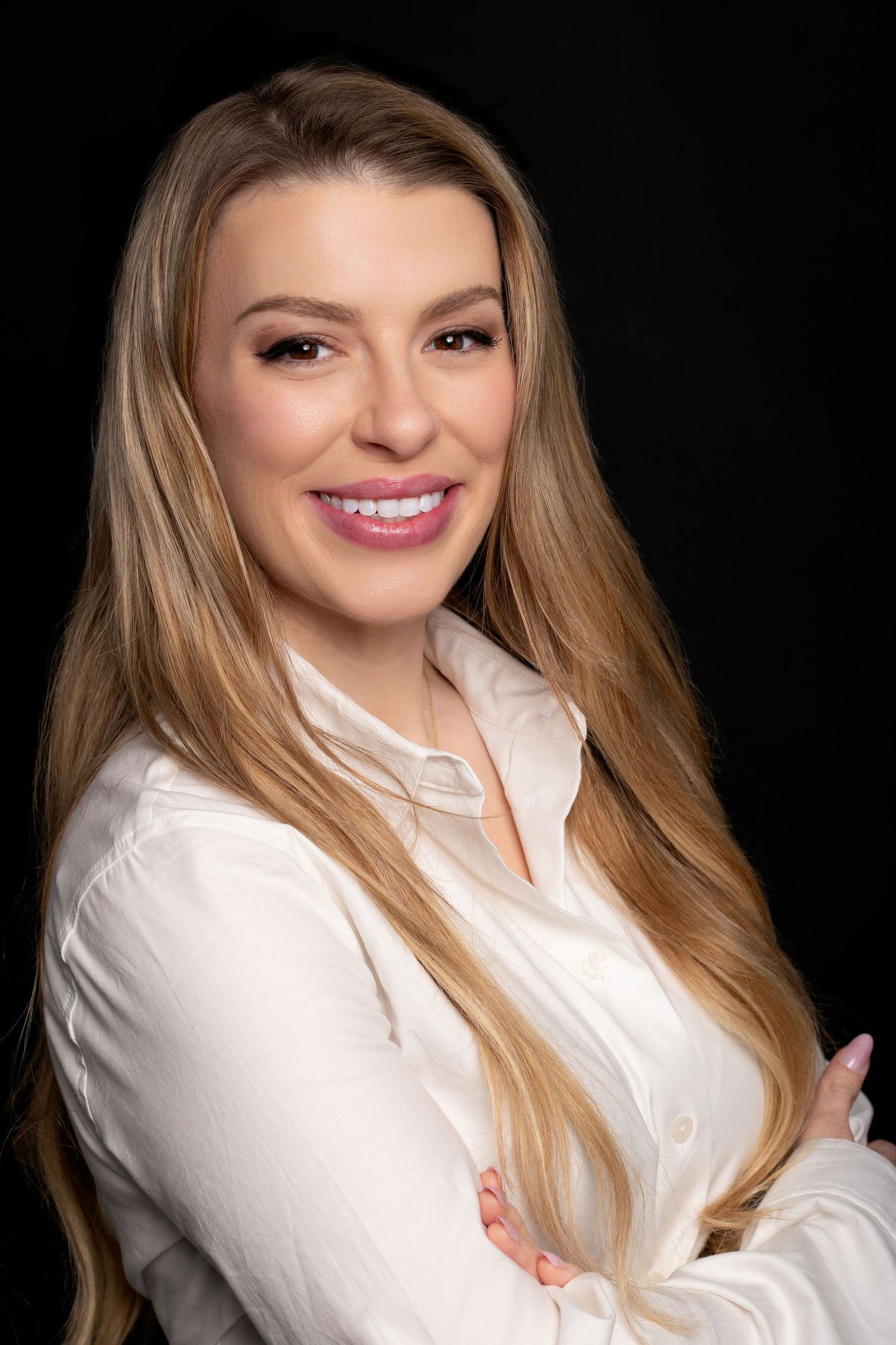 Blonde woman in white button-up shirt smiles with arms crossed in professional headshot on black background.