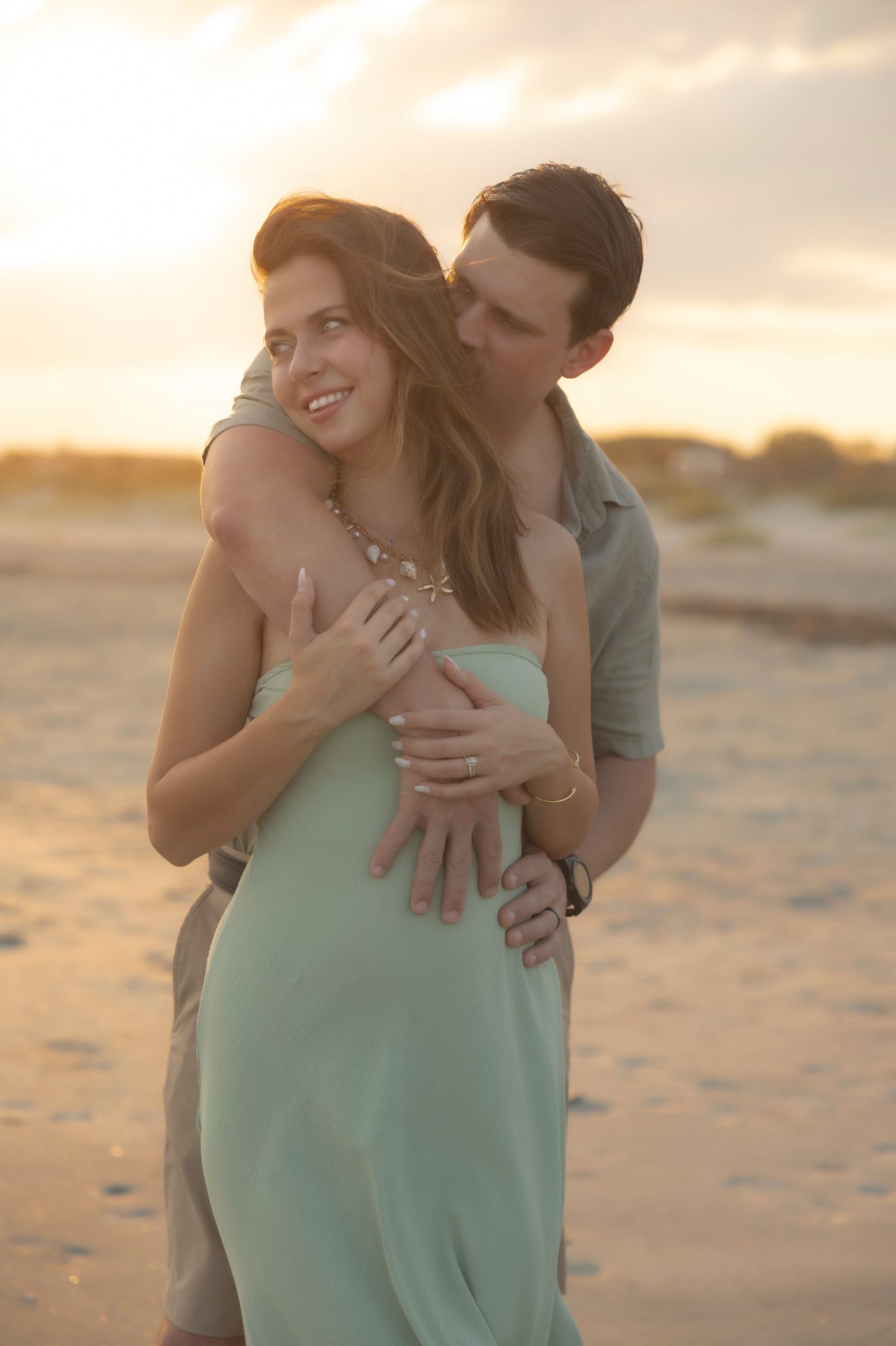 Couple embracing on beach at golden sunset, woman in mint dress smiling as man holds her from behind.