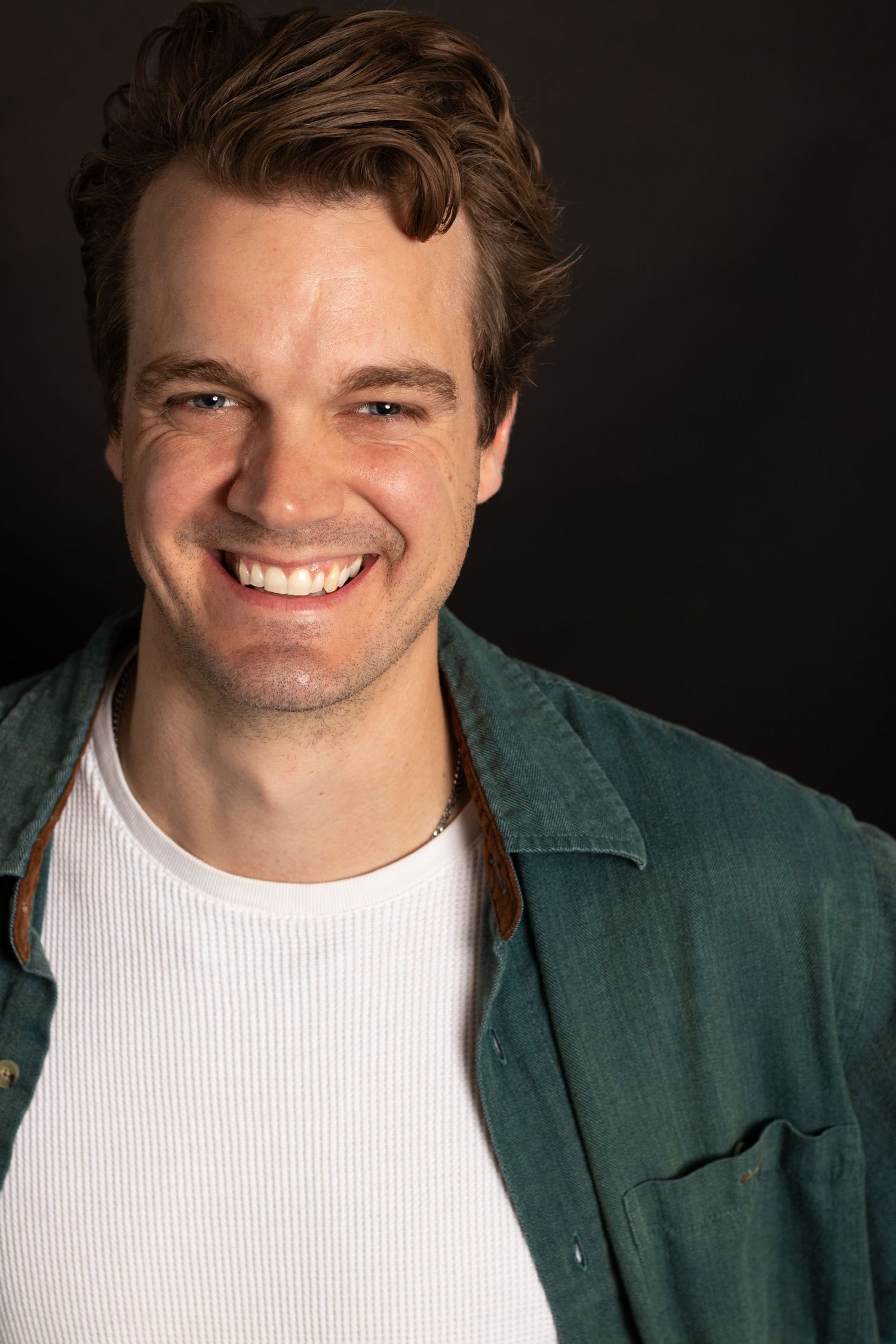 Smiling young man with brown hair wearing green jacket over white shirt against dark background.